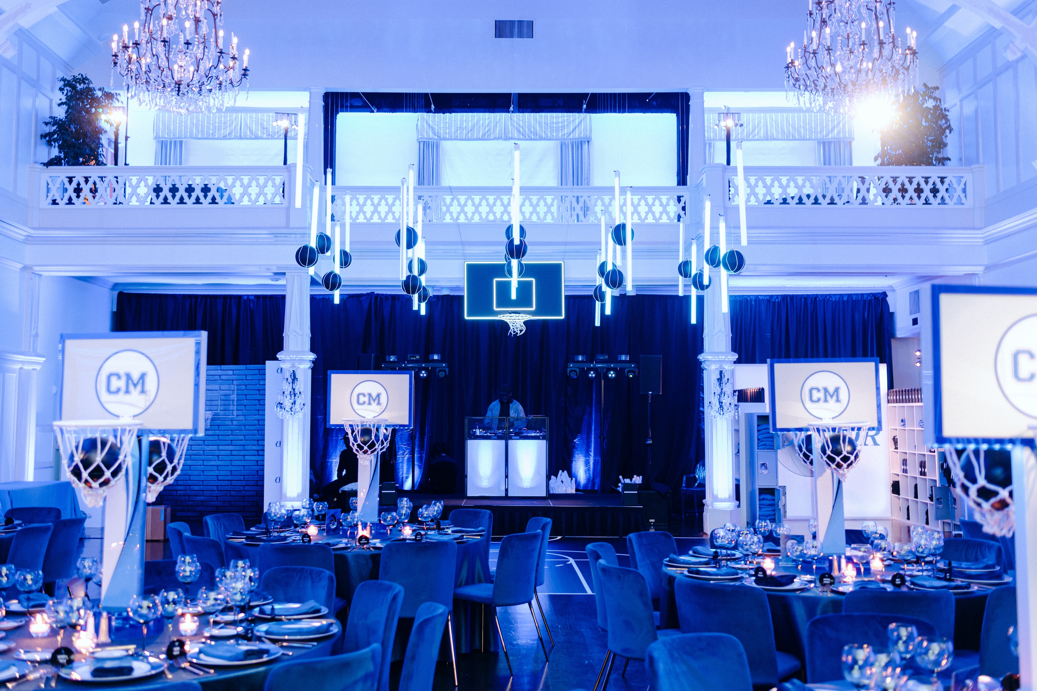 A lavish, blue-lit event space features elegant chandeliers, round tables set for dinner, and basketball-themed decorations, including a neon hoop.