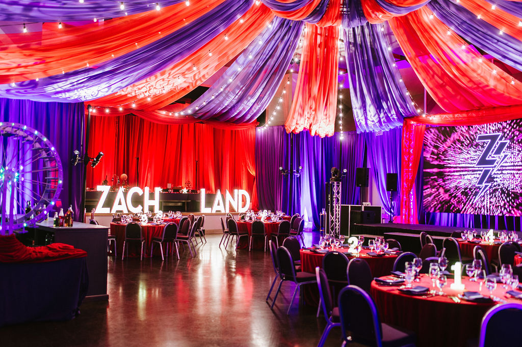 The image shows a vibrant event space adorned with red and purple drapes, glowing “ZACH LAND” letters, round tables with elegant settings, and a colorful stage backdrop.