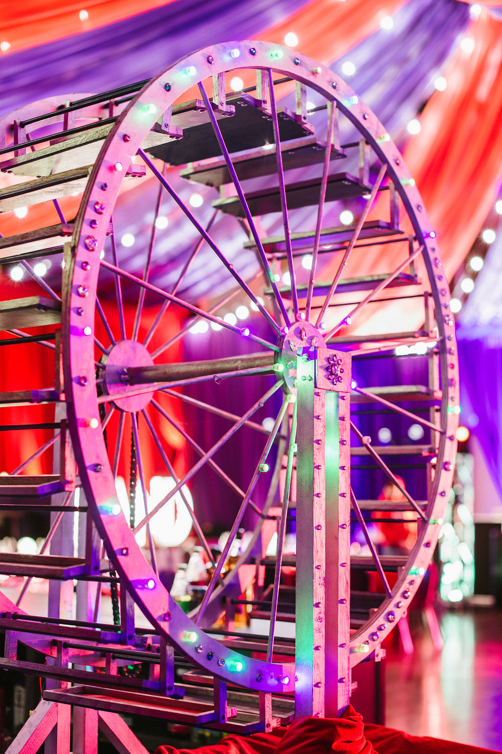 A brightly lit Ferris wheel structure with multicolored lights is set against vibrant red and purple draped fabric, creating a festive atmosphere.