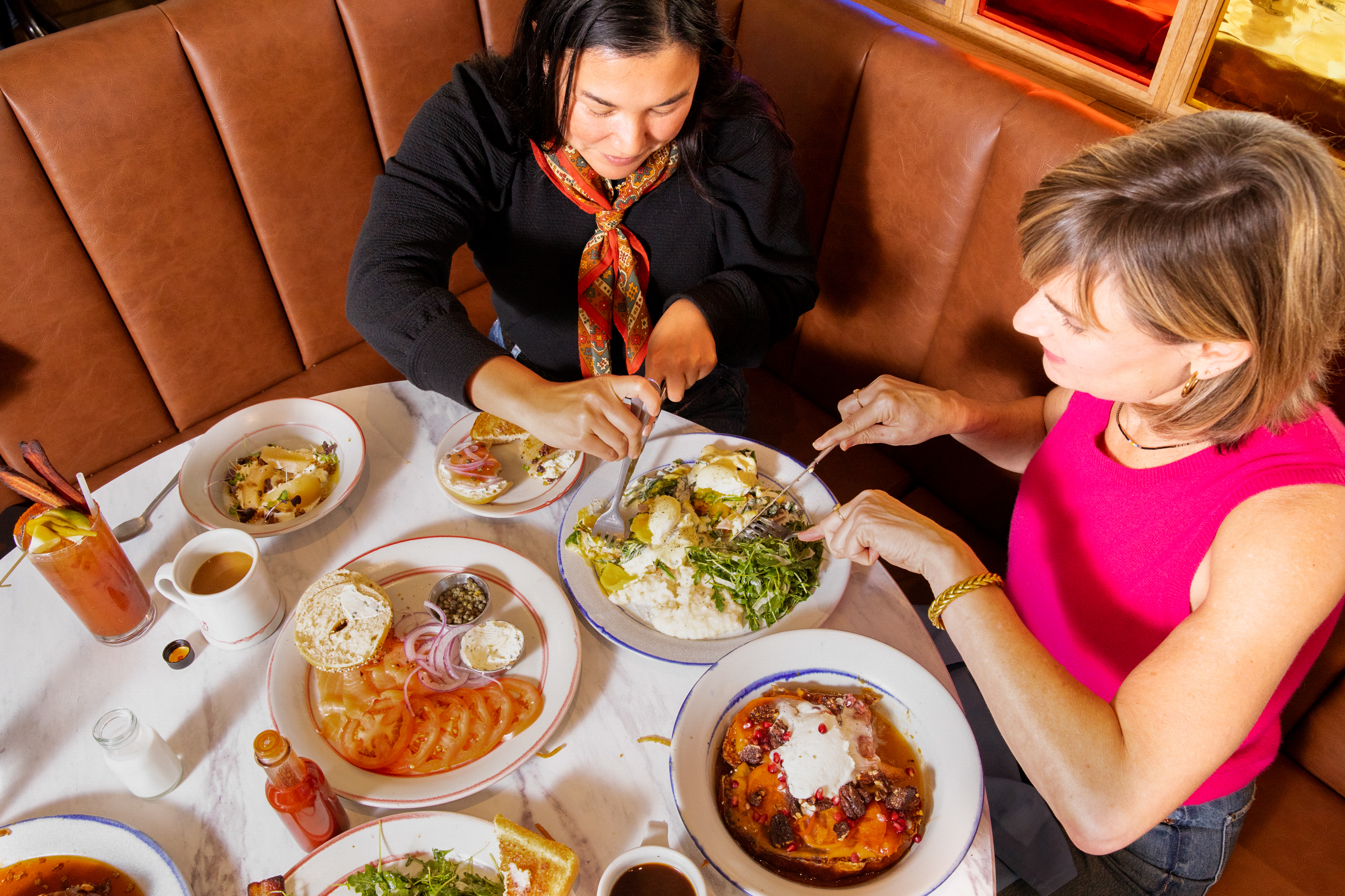 Two people are sitting at a table sharing a meal. Plates include salad, bagels with toppings, and a dish with fruit. Drinks include coffee and a cocktail.