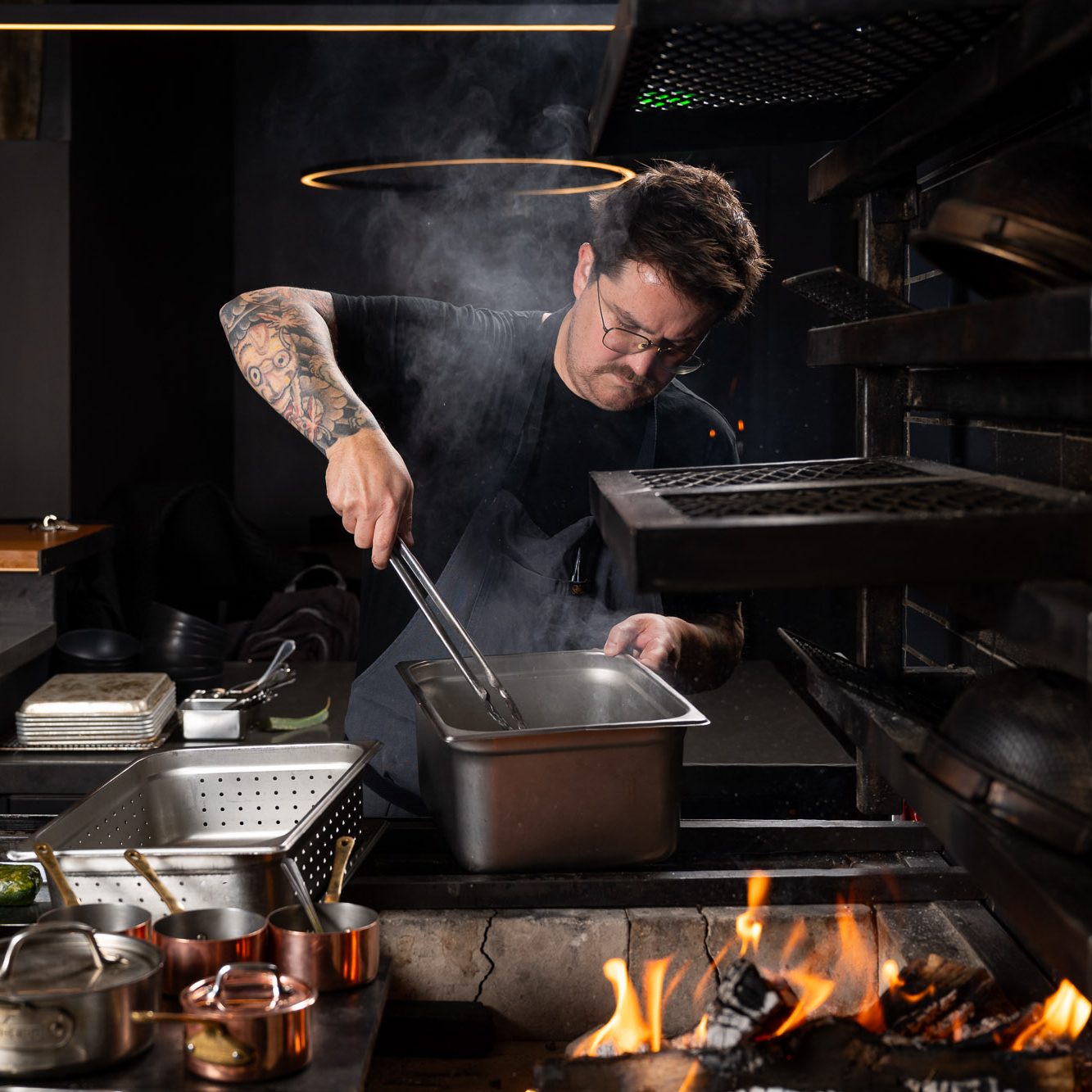 A chef with tattoos is focused on cooking at a smoky stove, using tongs over a metal pan while flames burn in the foreground and copper pots sit nearby.