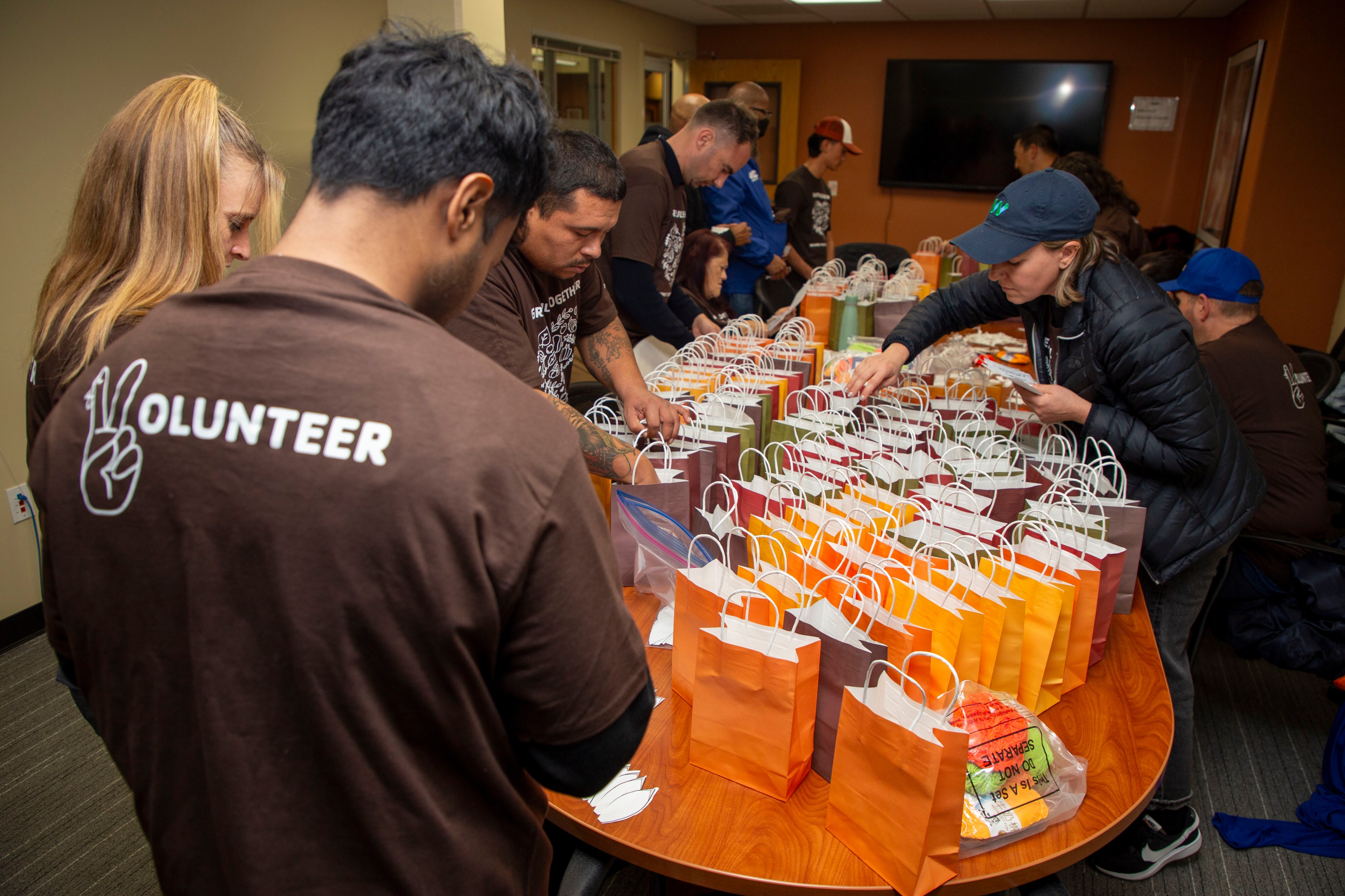 Volunteers in brown shirts are organizing colorful gift bags on a table, filled with various items, in a room with a TV on the wall.