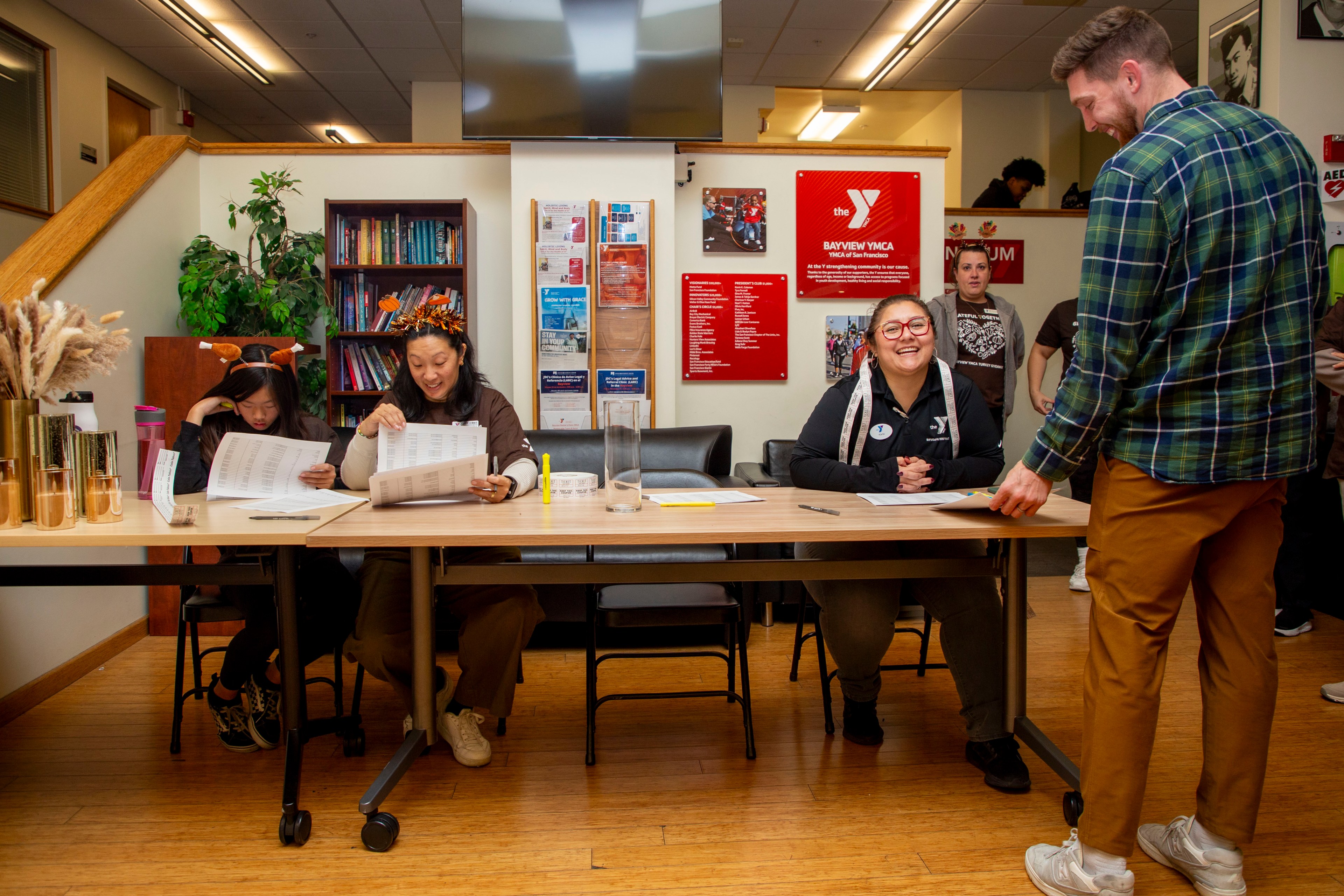 Three people sit behind a table with papers, smiling and interacting with a standing man. The room is decorated with posters and plants, creating a welcoming atmosphere.