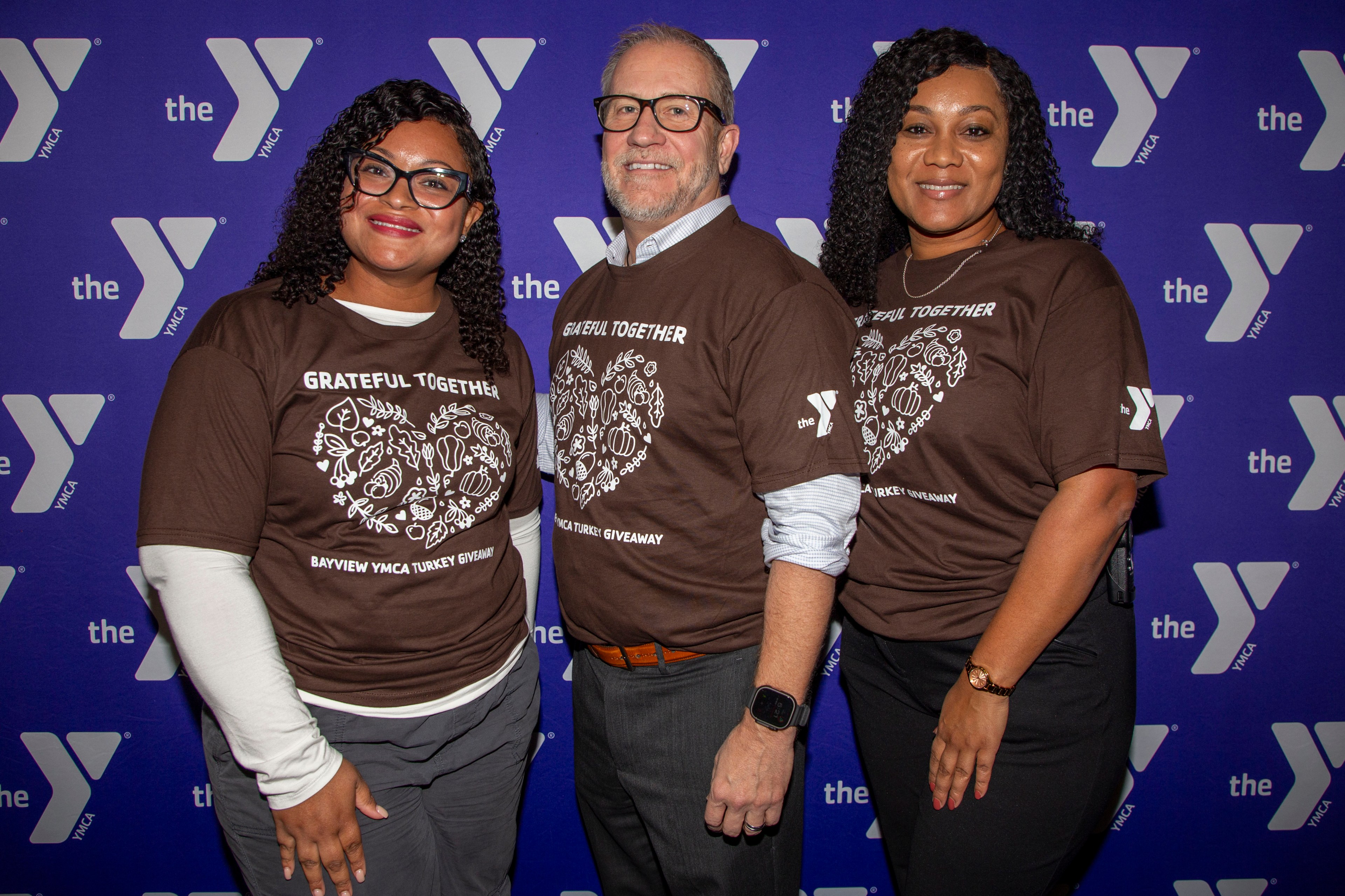 Three people smile at the camera, wearing matching brown shirts with &quot;Grateful Together&quot; on them, in front of a YMCA backdrop.