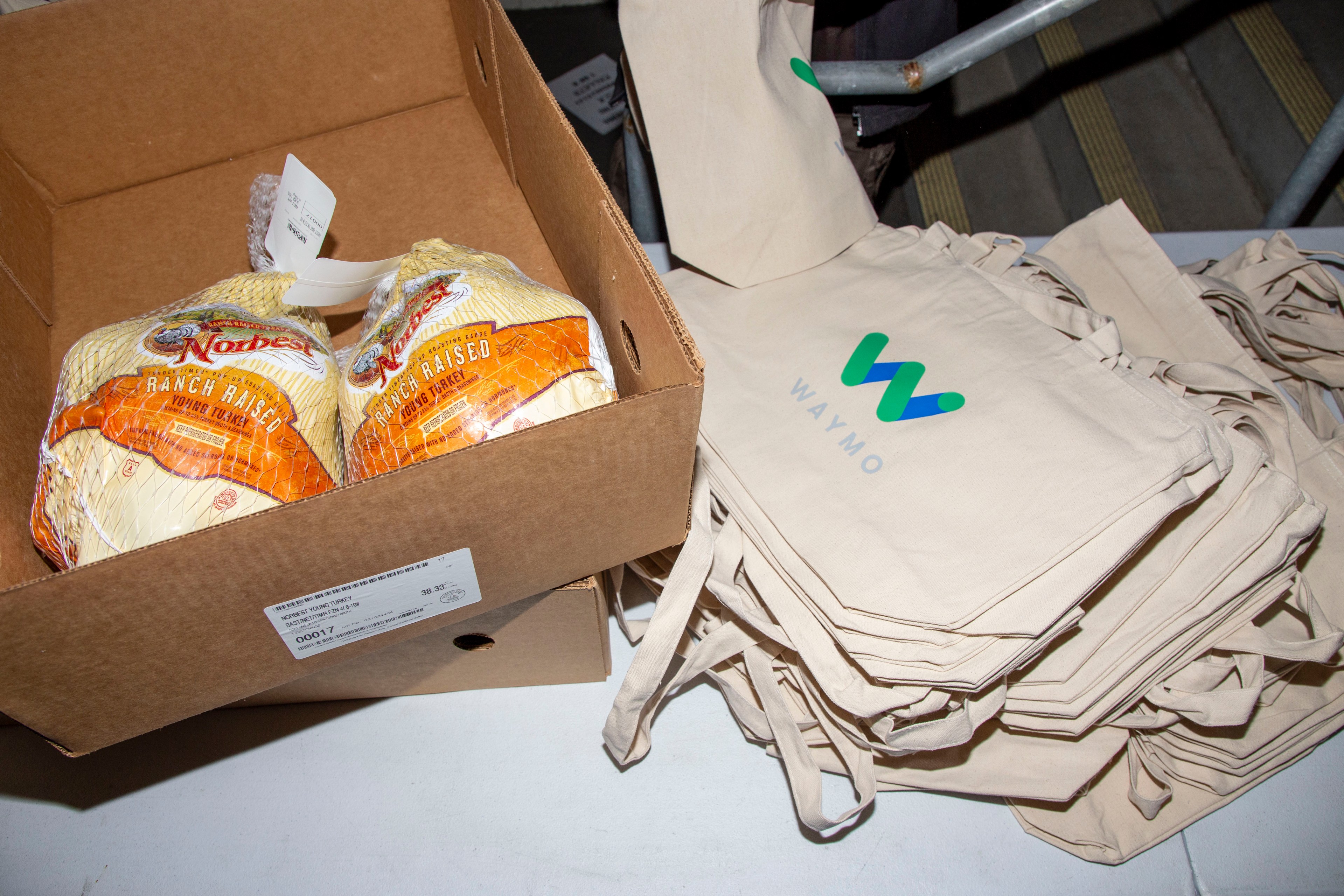 The image shows a cardboard box with two packaged turkeys next to a stack of beige tote bags with the Waymo logo on them.