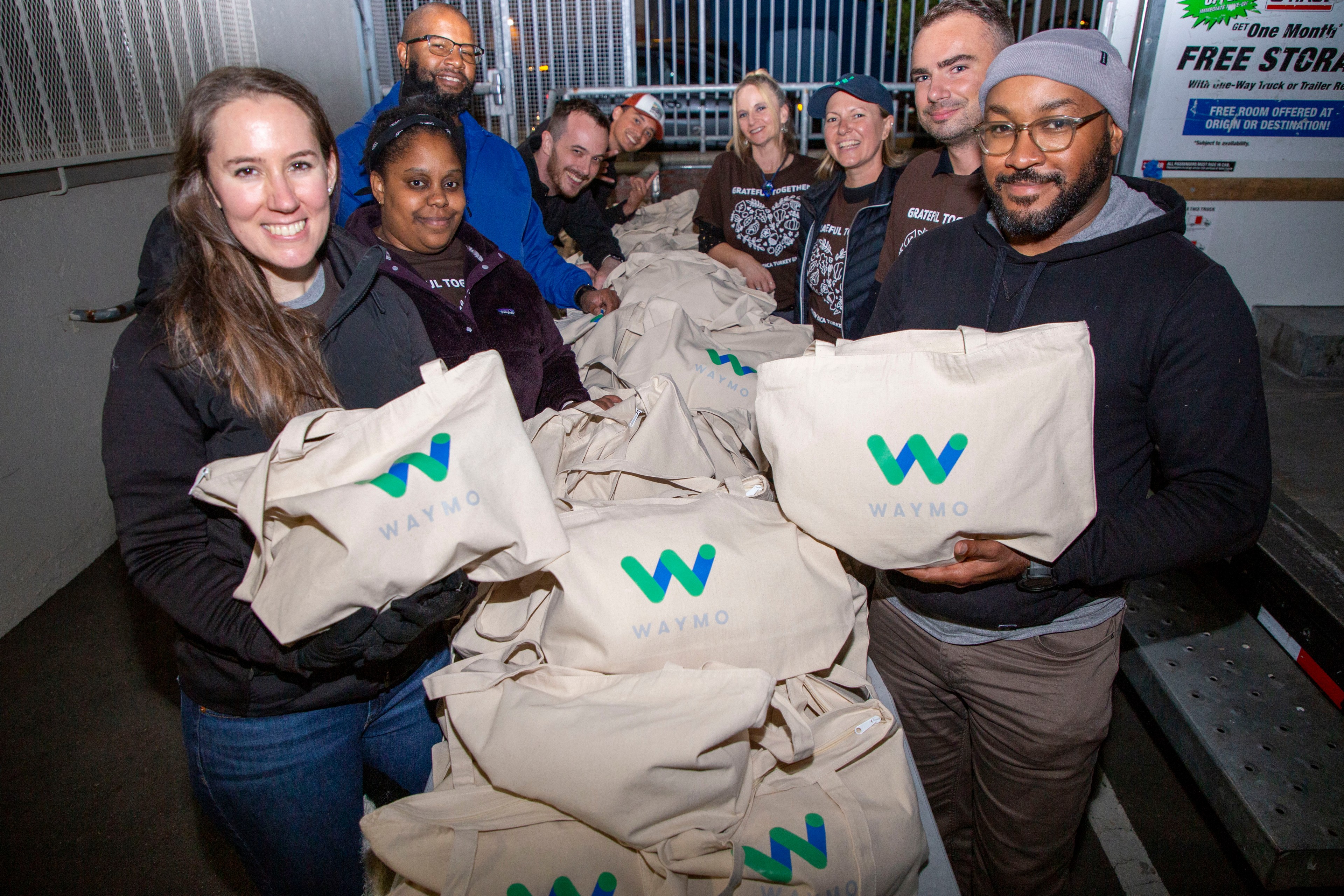 A group of people smiling and holding beige bags with the &quot;Waymo&quot; logo. They appear to be at a distribution event, surrounded by many similar bags.