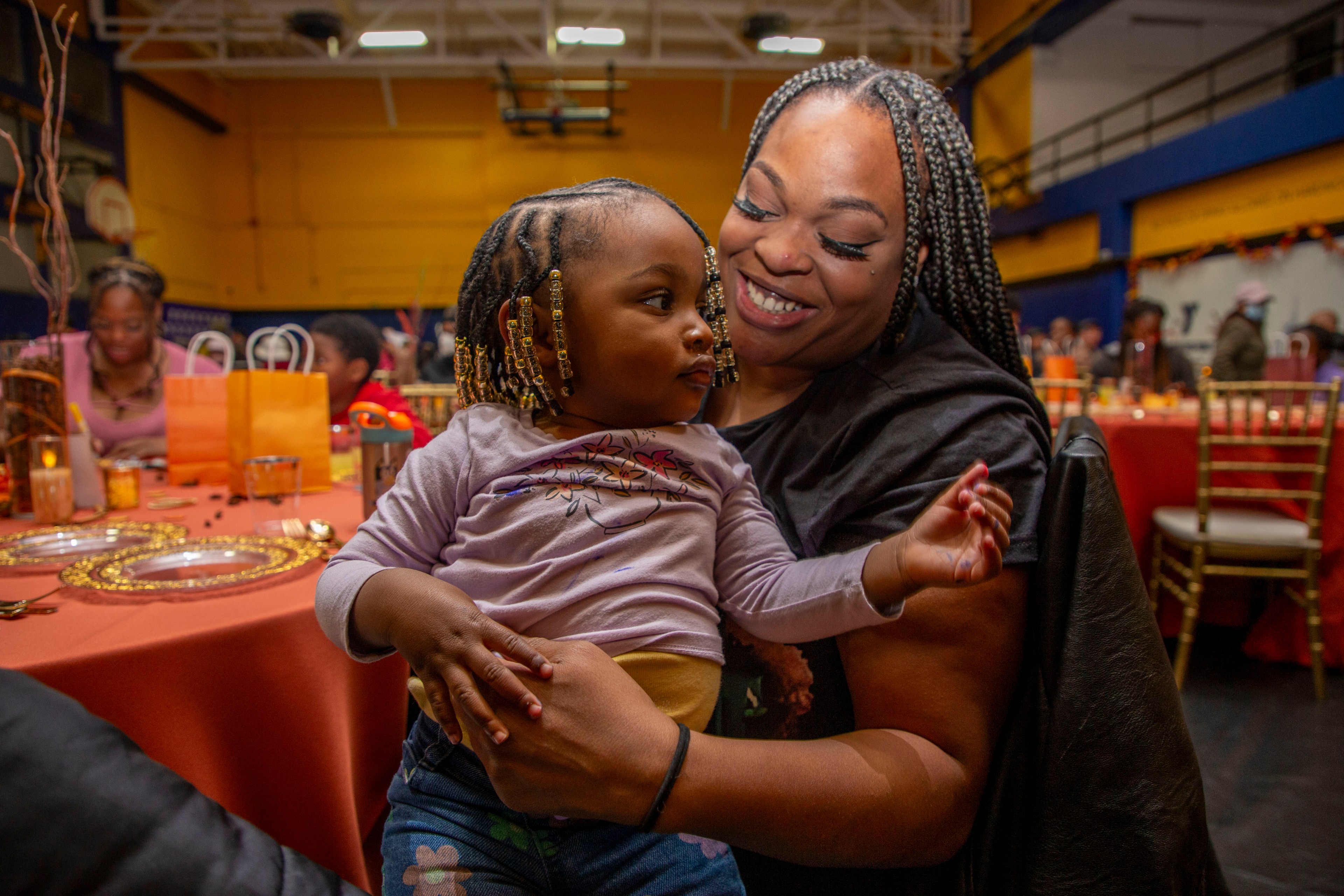 A smiling woman holds a young child with braided hair in a lively indoor setting with decorated tables and orange-themed gifts in the background.