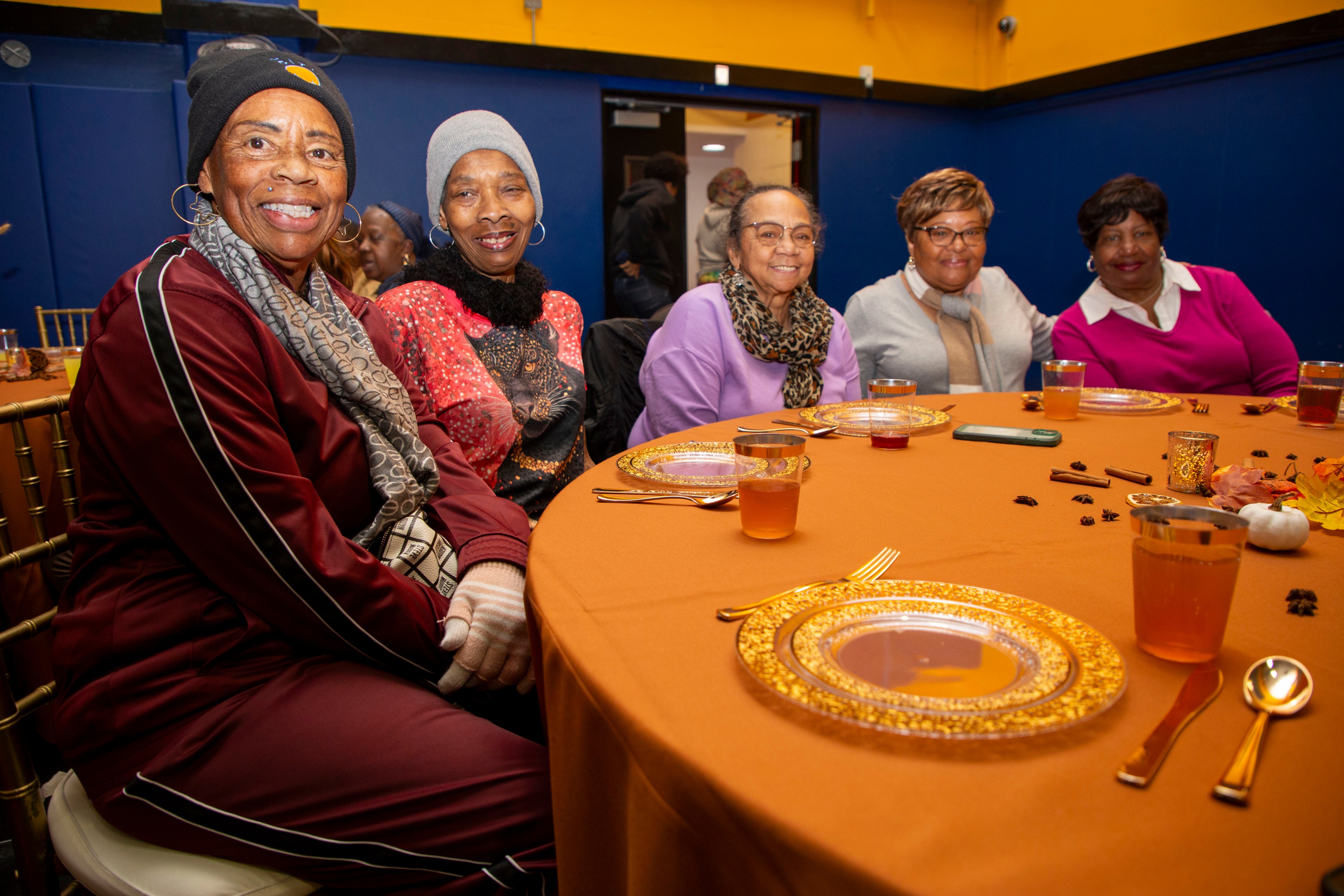 Five women sit smiling at a round table with orange tablecloth and decorative plates. They appear to be enjoying a gathering in a warmly lit room.