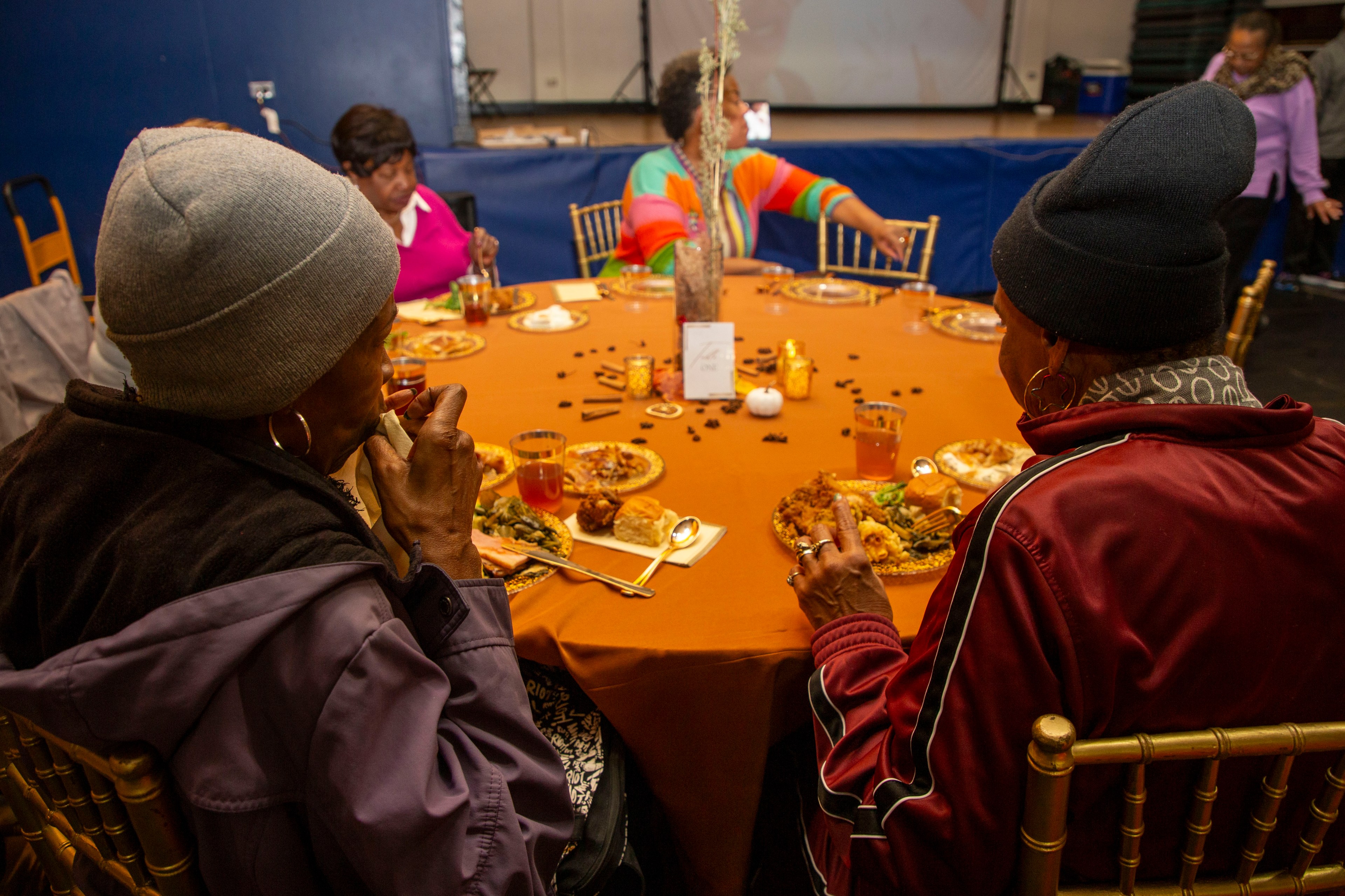 Several people are sitting around a round table with an orange tablecloth, enjoying a meal. The table is set with plates, drinks, and autumn-themed decor.