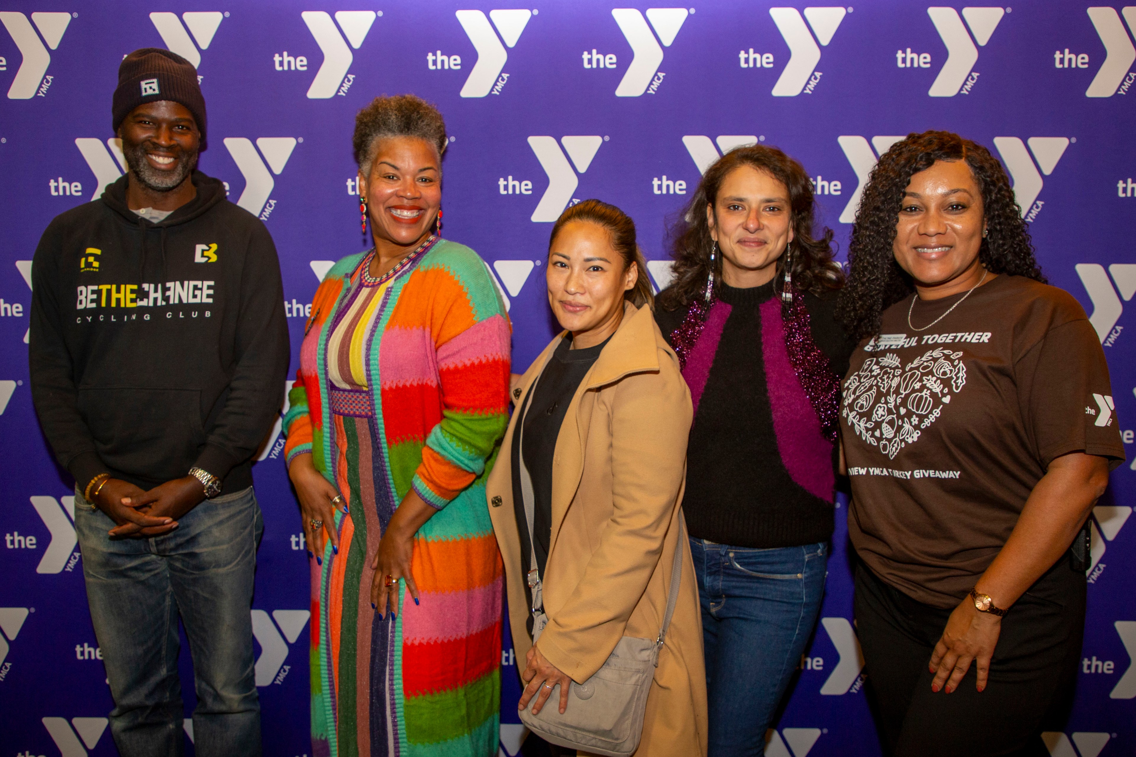 Five people stand smiling in front of a YMCA backdrop, with diverse styles ranging from a colorful coat to a brown t-shirt, capturing a sense of togetherness.