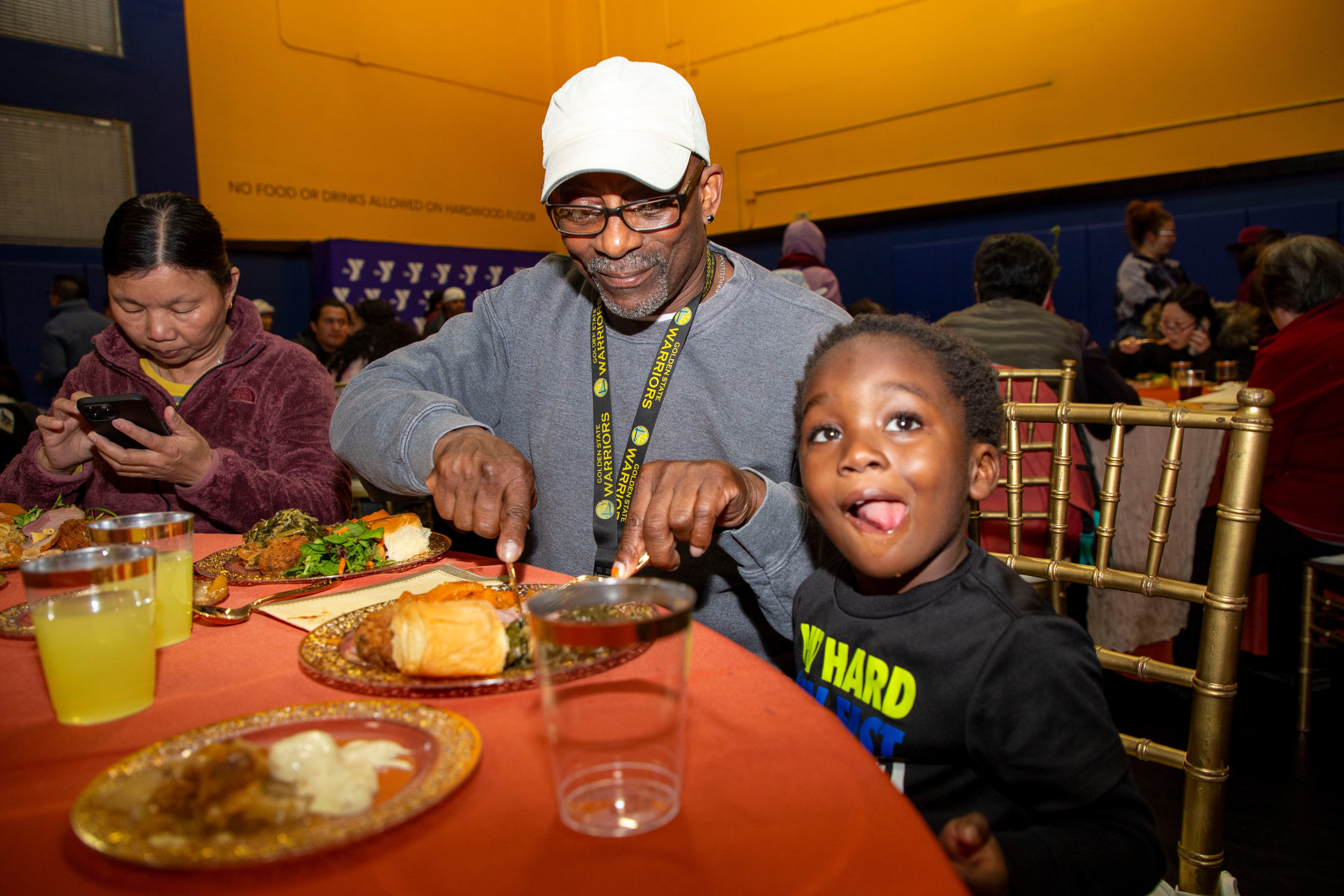 A man in a white cap and glasses smiles while cutting food on his plate. A child beside him playfully sticks out their tongue, sitting at a decorated table.