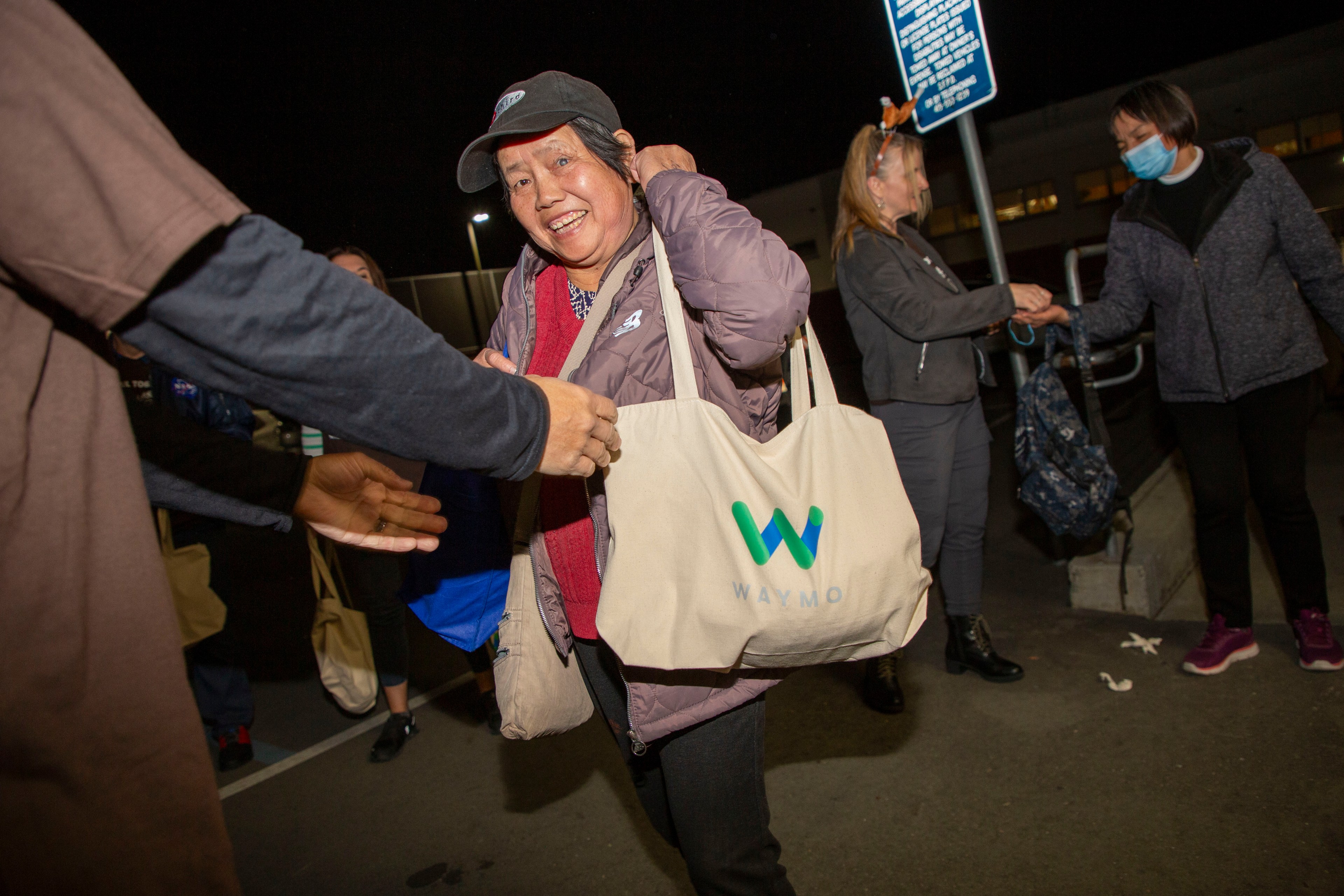 A smiling woman wearing a cap holds a Waymo tote bag among a group of people interacting at night, some of whom are exchanging handshakes.