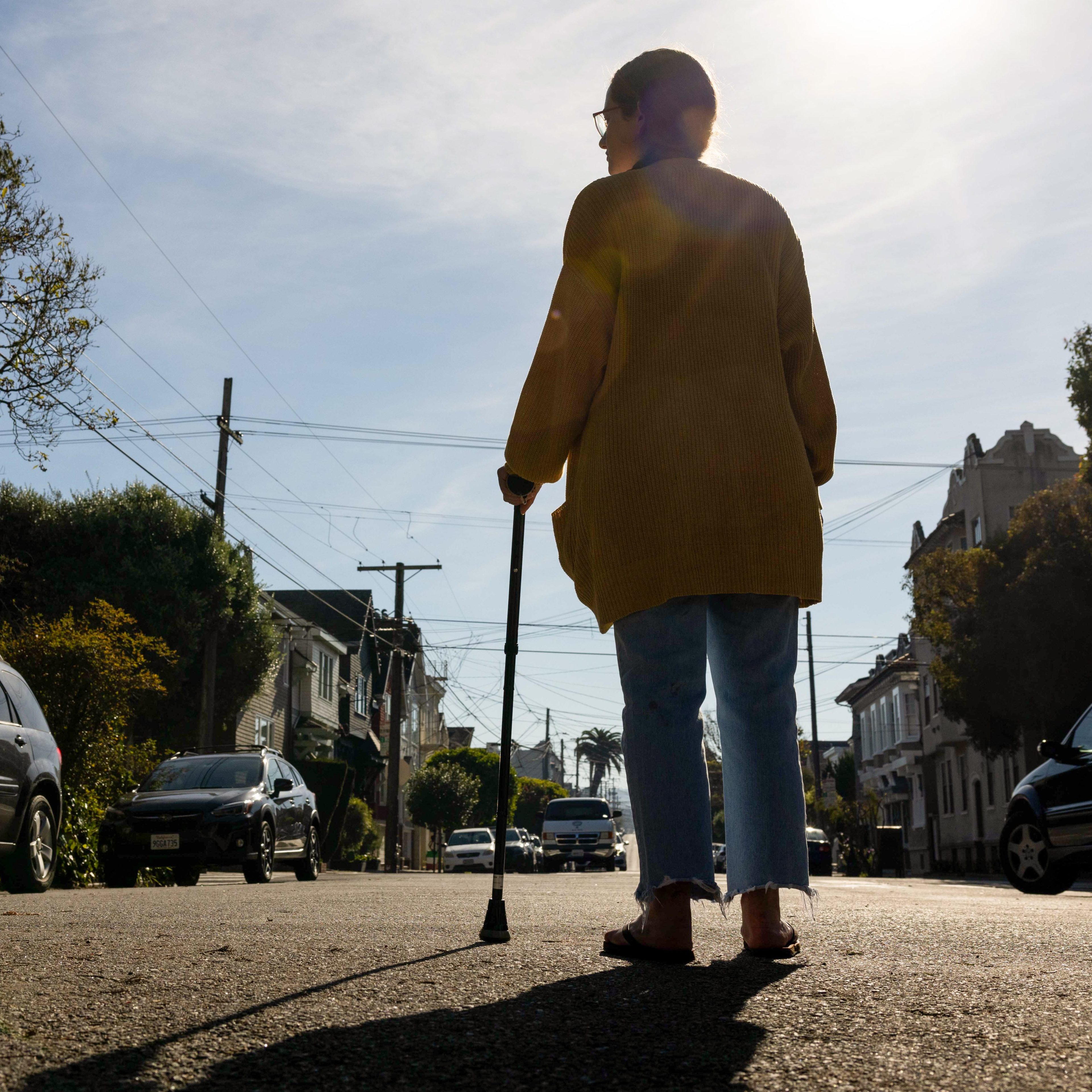 A person wearing a yellow sweater and blue jeans, using a cane, stands in the middle of a sunlit street with cars parked on both sides.