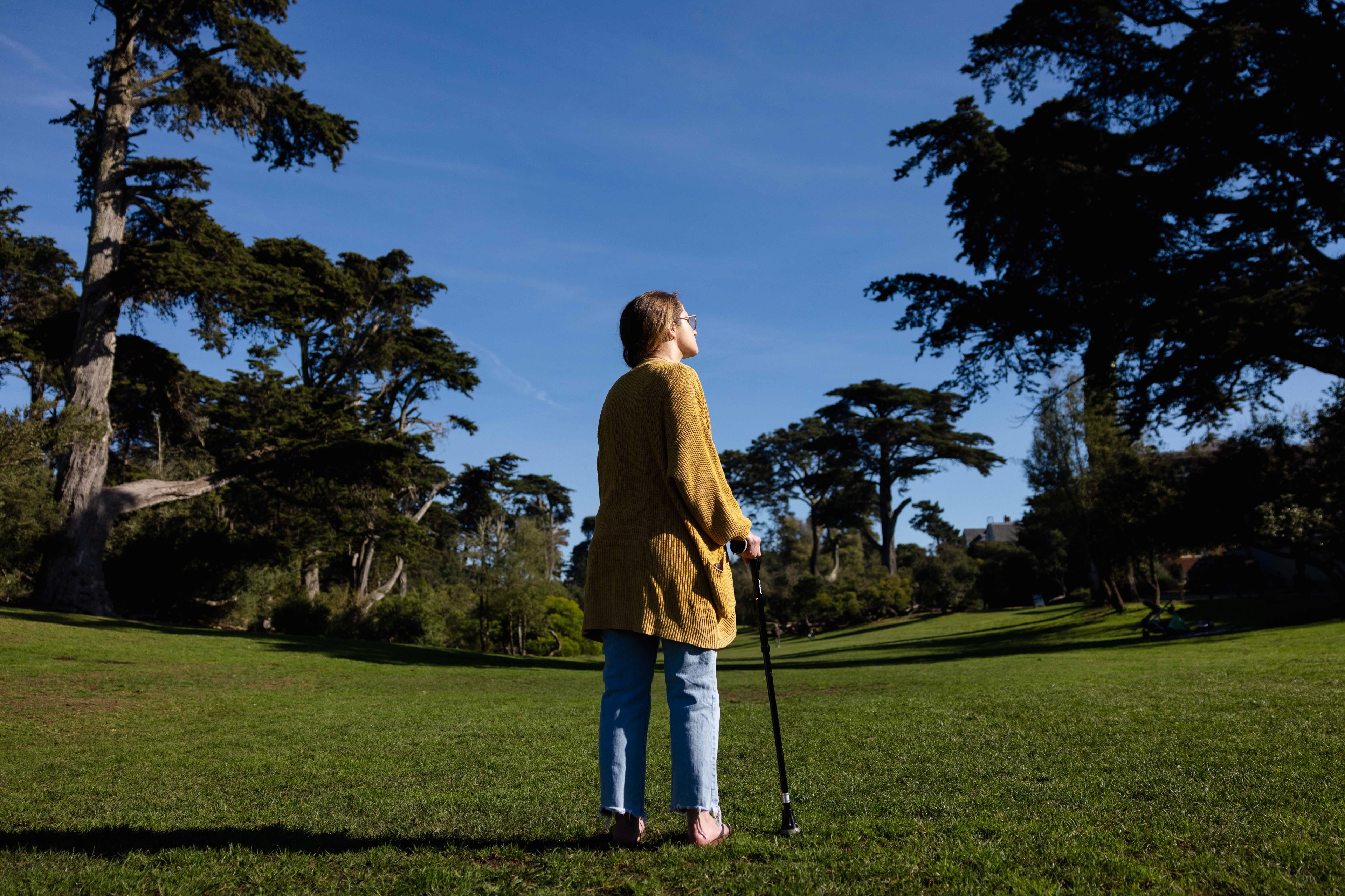 A person stands on a grassy field using a cane, facing away. They're wearing a mustard sweater and jeans, with tall trees and a clear blue sky in the background.