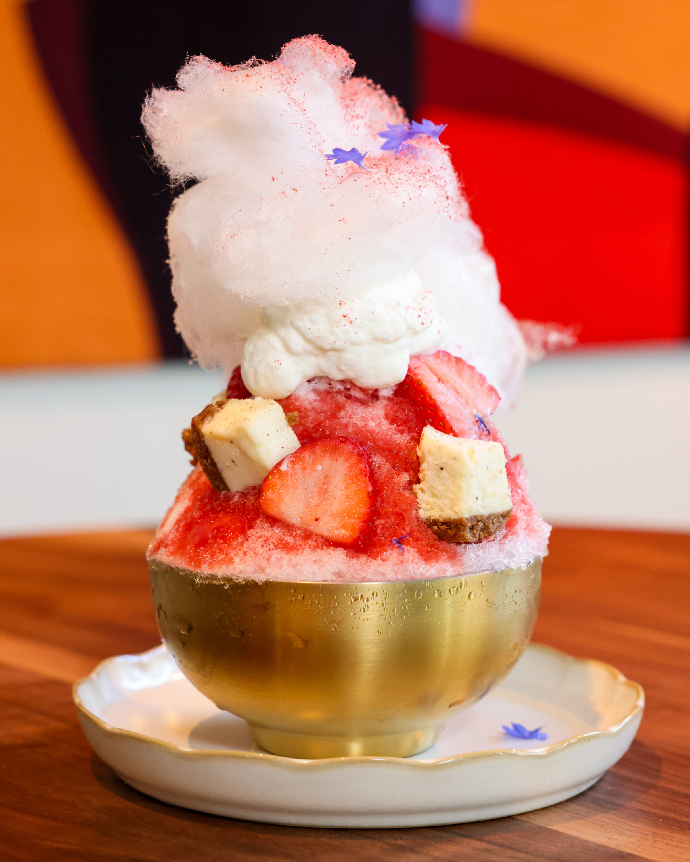 A golden bowl holds a layered dessert with pink shaved ice, strawberries, cream, and cubes of sponge cake, topped with fluffy cotton candy and blue flower petals.