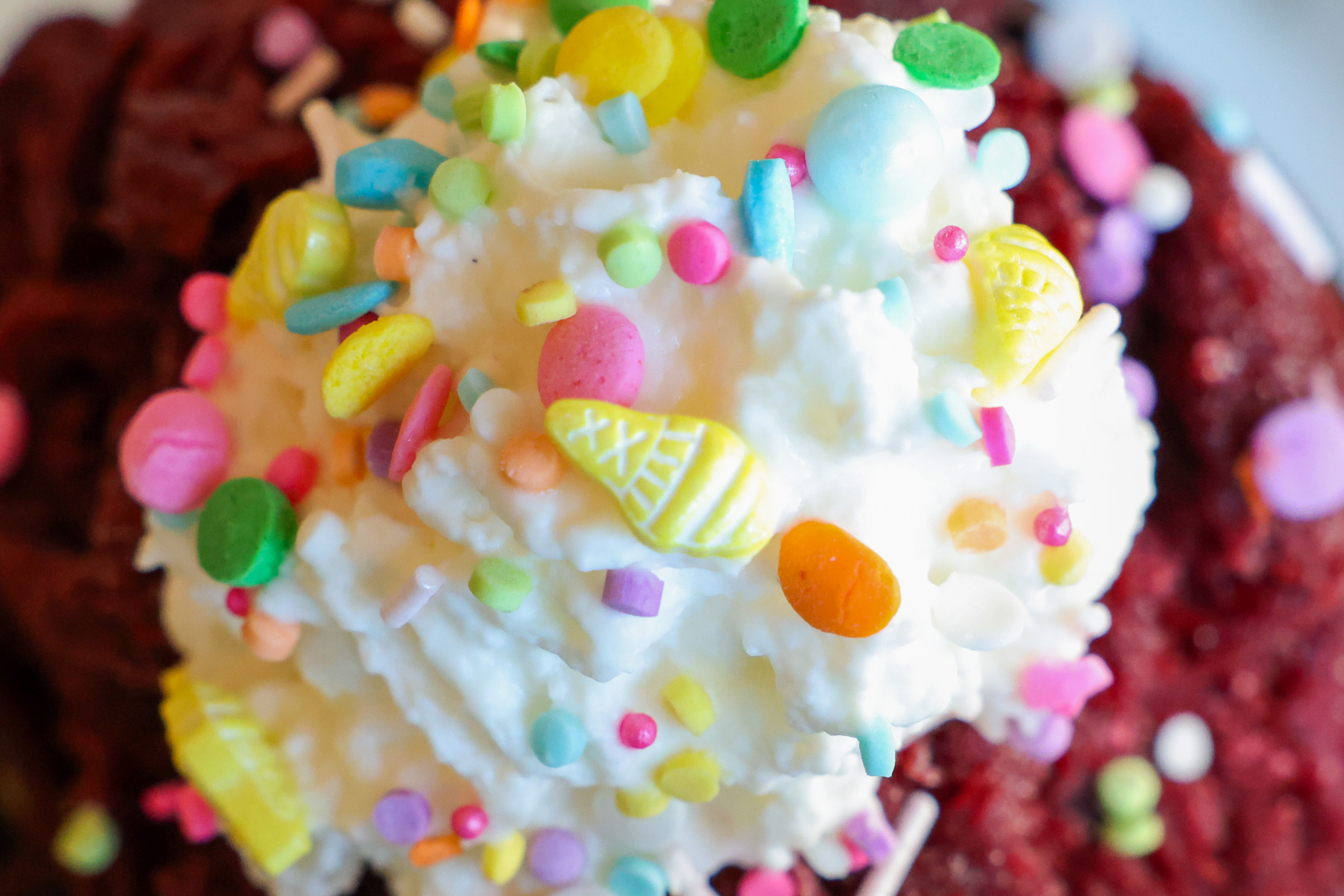 The image features a swirl of whipped cream topped with colorful sprinkles and small candy pieces, set upon a deep red dessert.