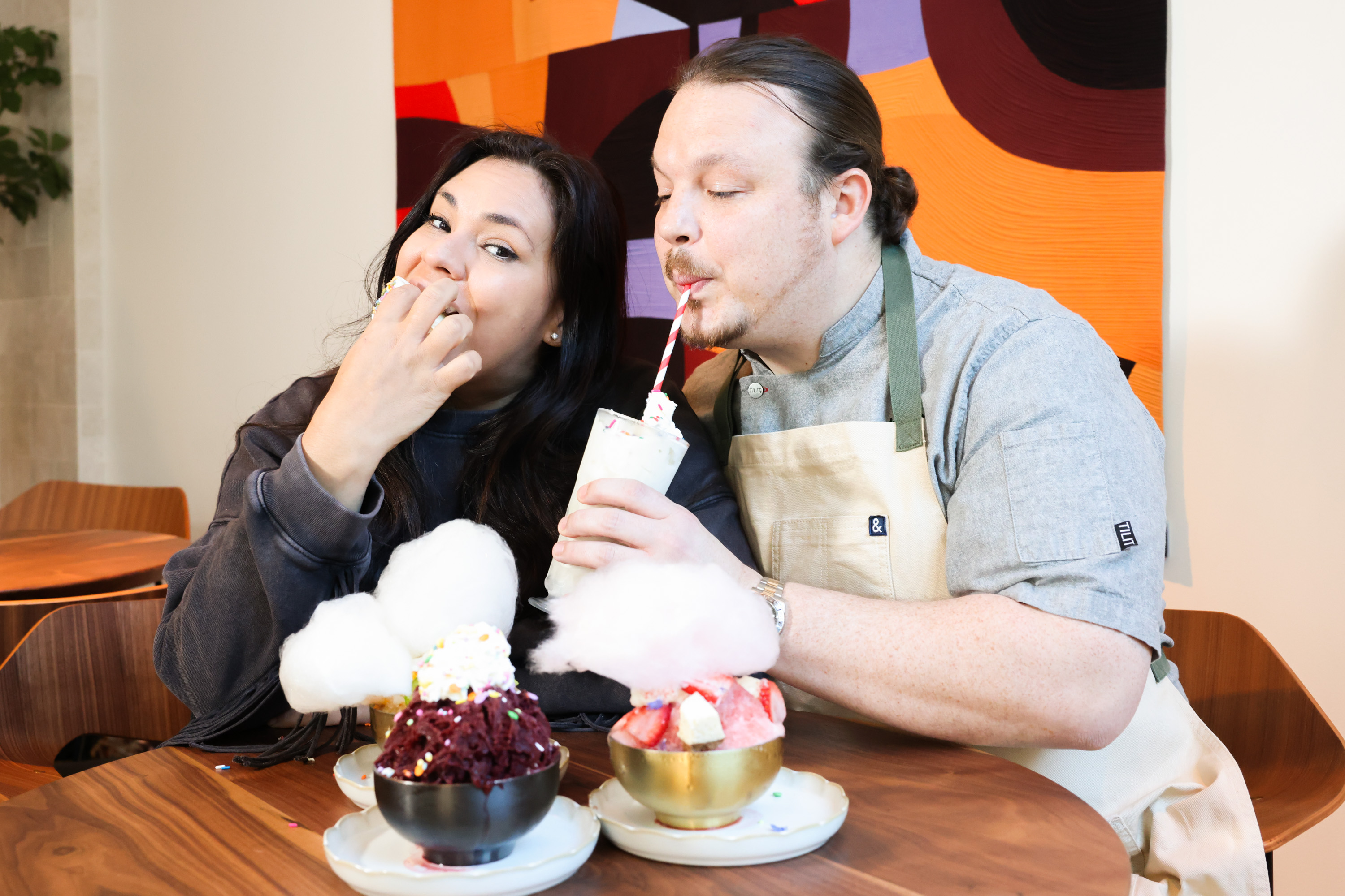 Two people enjoy desserts, with one sipping from a milkshake and the other eating with fingers. Colorful bowls of treats and a bold abstract art piece are present.