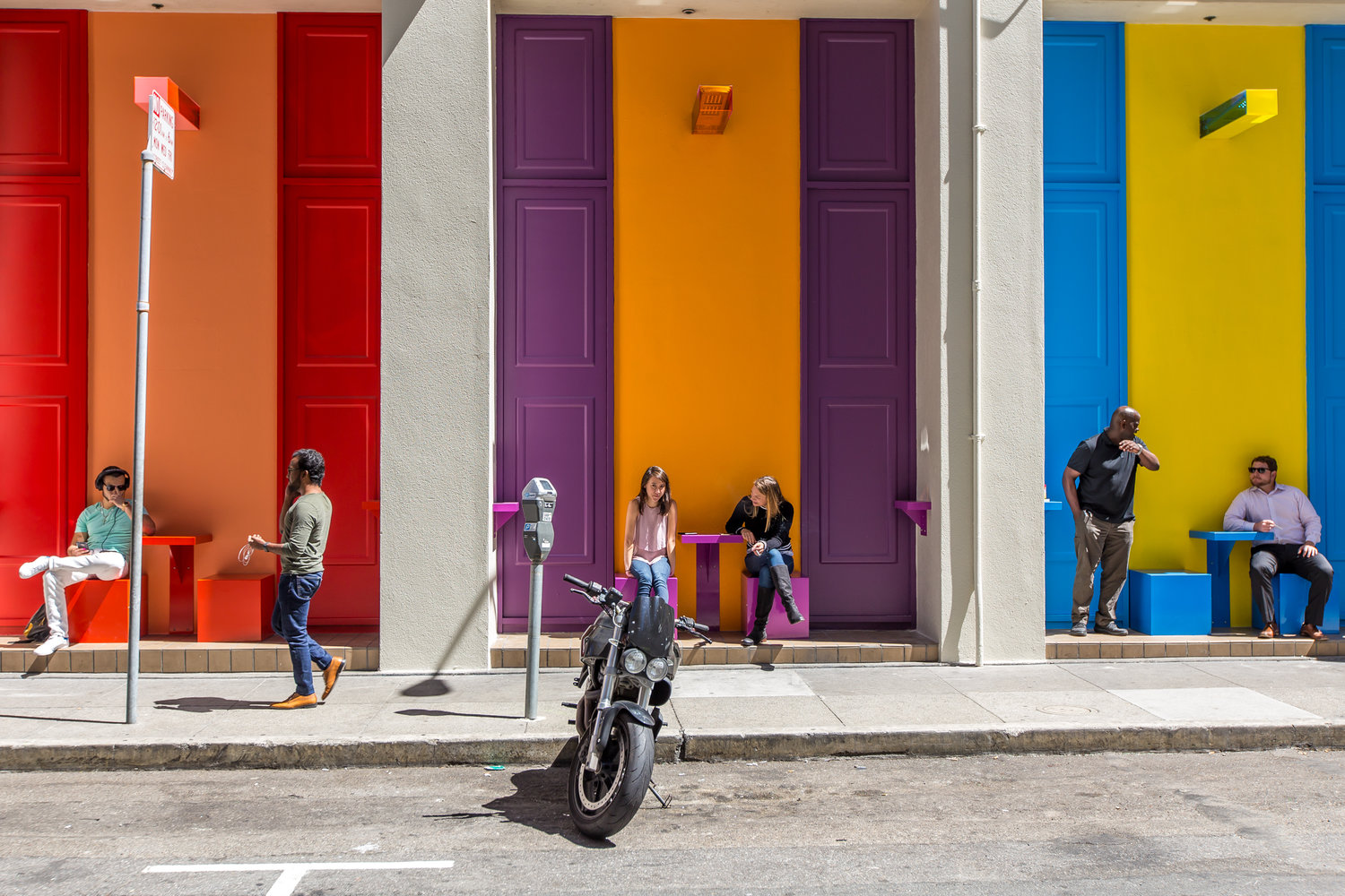 The image shows people sitting and interacting against a vibrant wall with colorful squares: red, yellow, and blue. A motorcycle is parked on the street.