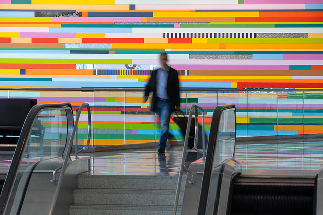 A person walks down steps between escalators, in front of a vibrant wall with colorful horizontal stripes in an indoor setting.