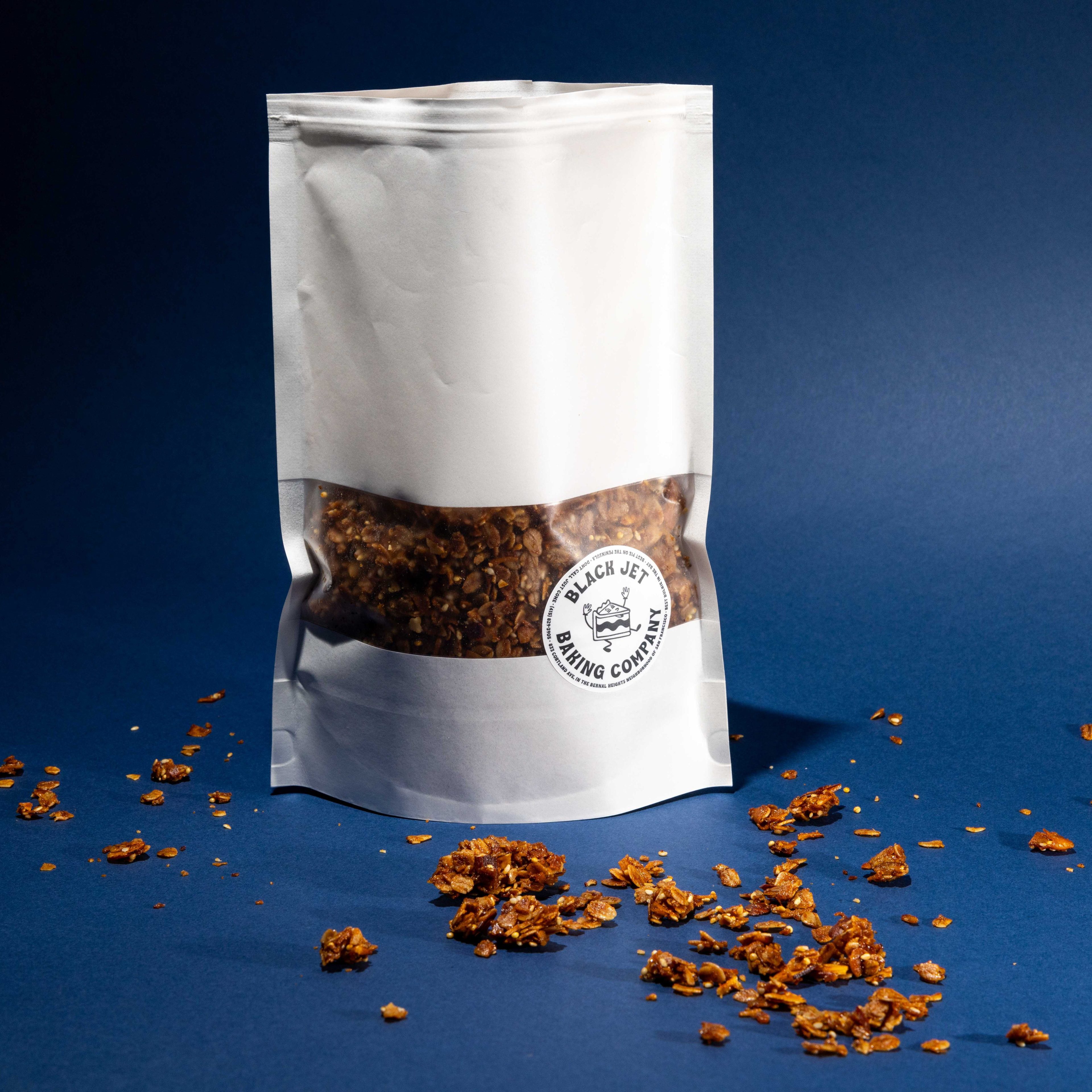 A white stand-up pouch of granola with a see-through window and a Black Jett Baking Company logo is on a blue surface, surrounded by scattered granola pieces.