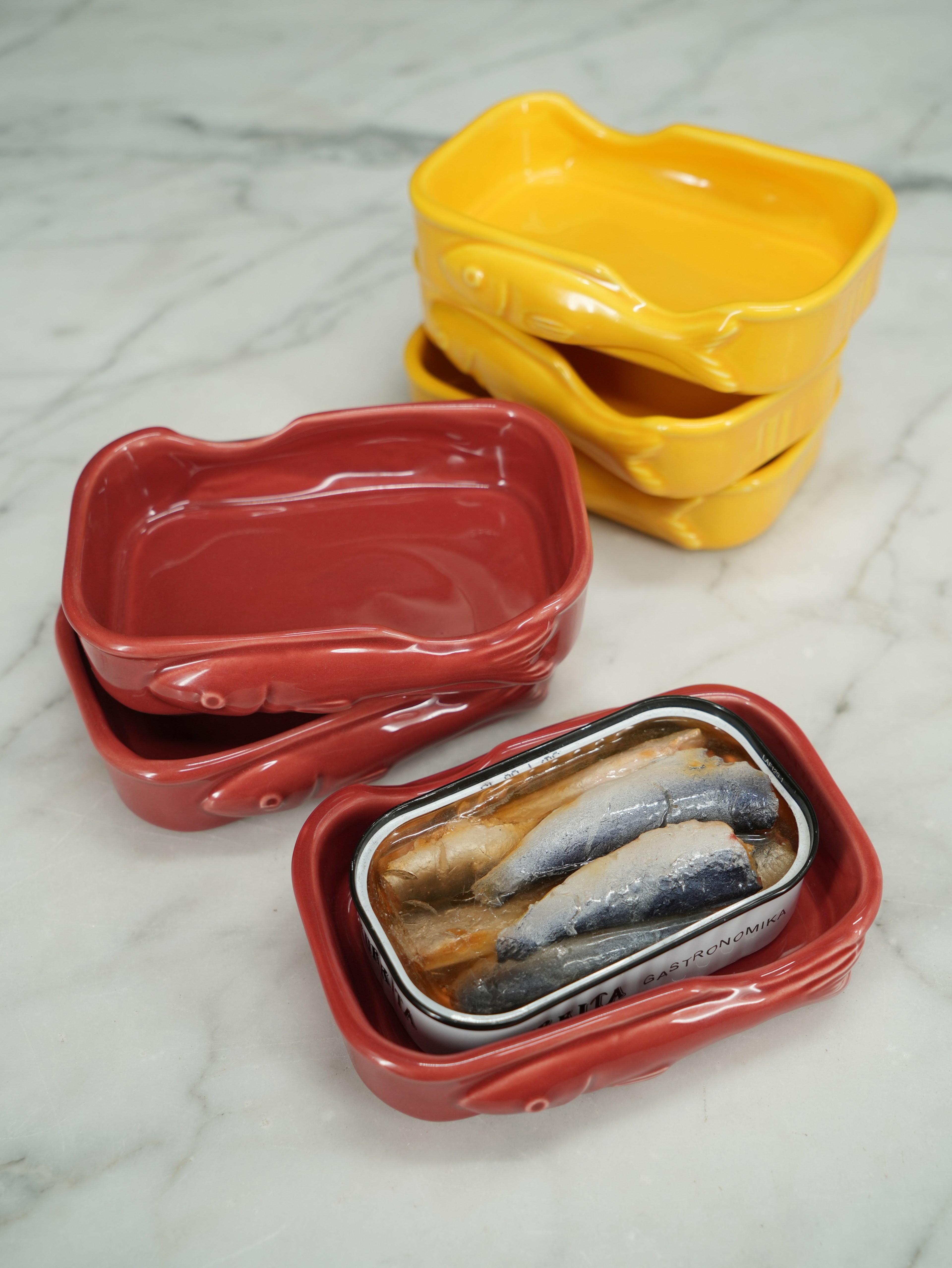 The image shows two stacked red fish-shaped dishes, two yellow ones stacked behind, and a tin of sardines placed in an open red dish on a marble surface.