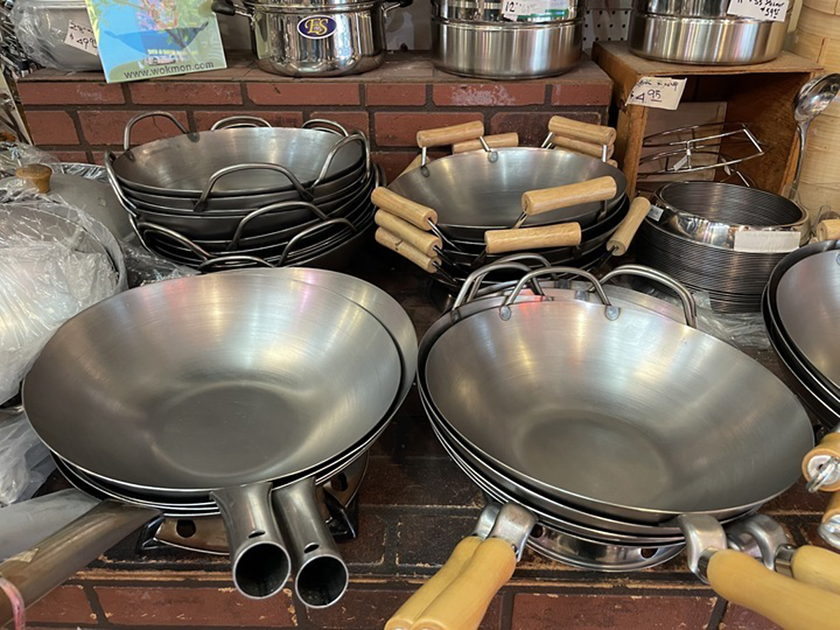 The image shows stacks of metal woks with wooden and metal handles on display in a store, surrounded by other cookware items against a brick wall backdrop.