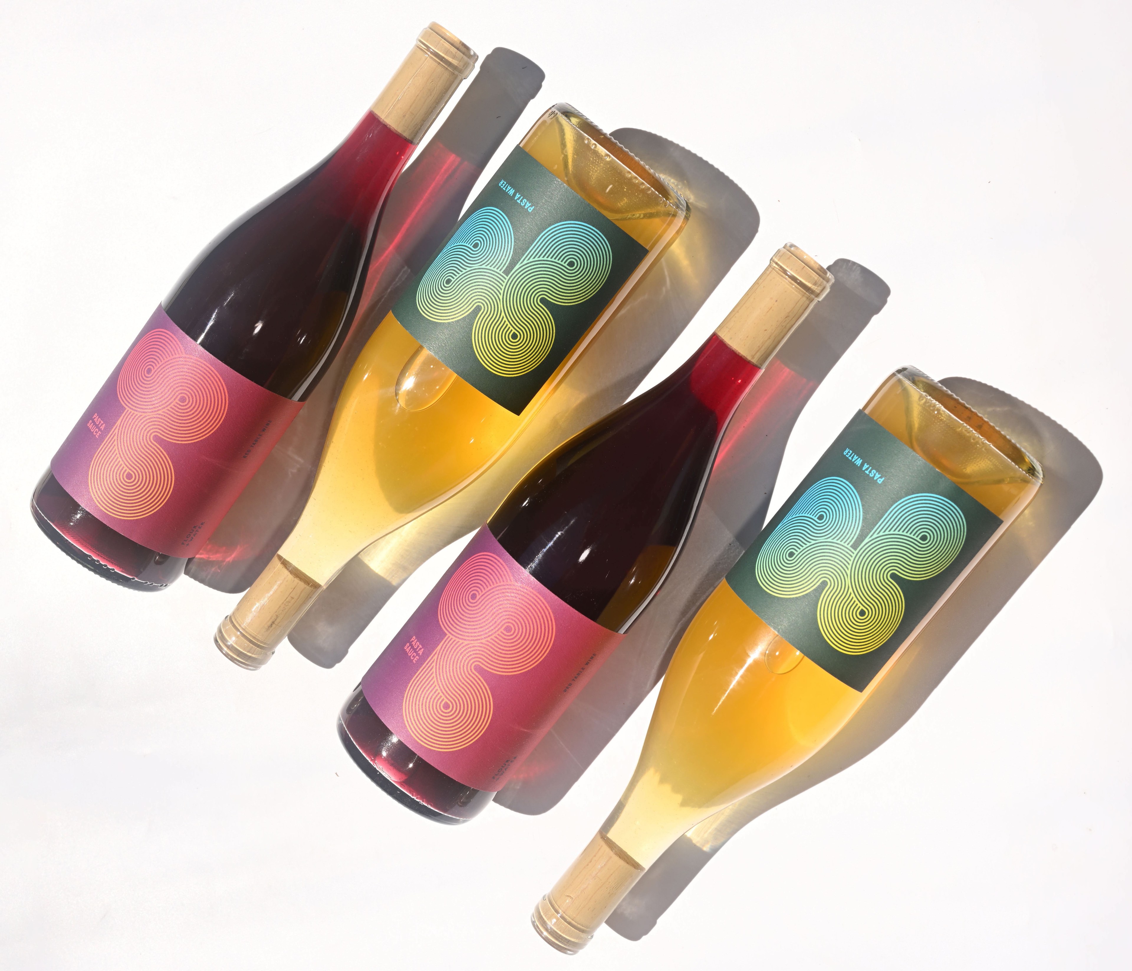 The image shows four wine bottles arranged diagonally. Two bottles have red labels with pink swirl designs, and two have teal labels with green swirl designs.