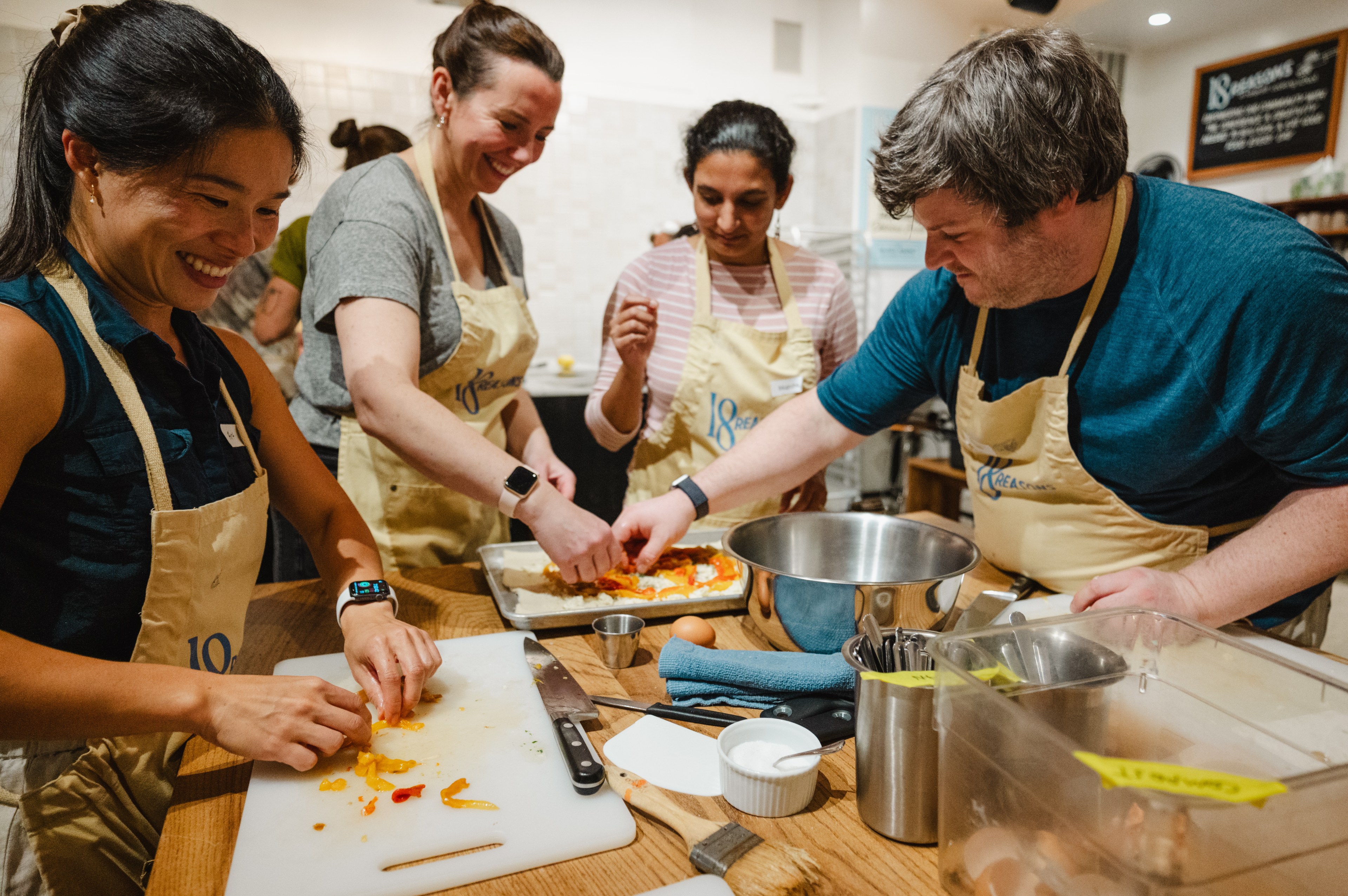 A group of four people wearing aprons are joyfully preparing food together, chopping and arranging ingredients on a kitchen counter.