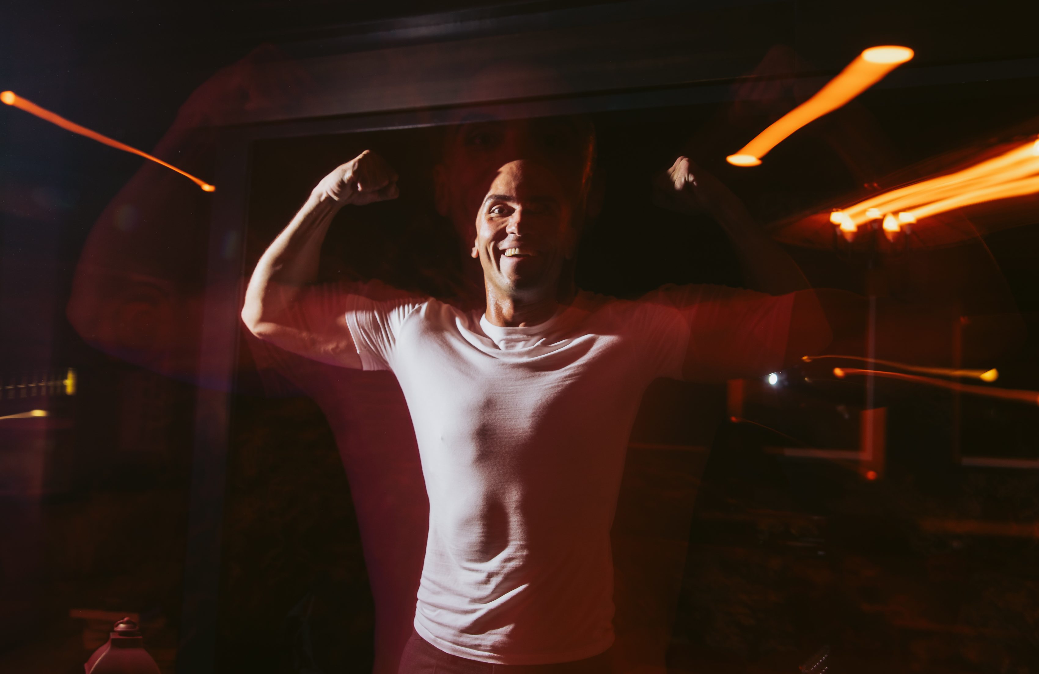 A person in a white shirt is flexing their muscles, with a smile, against a dark background. Light streaks create a dynamic, blurred effect.