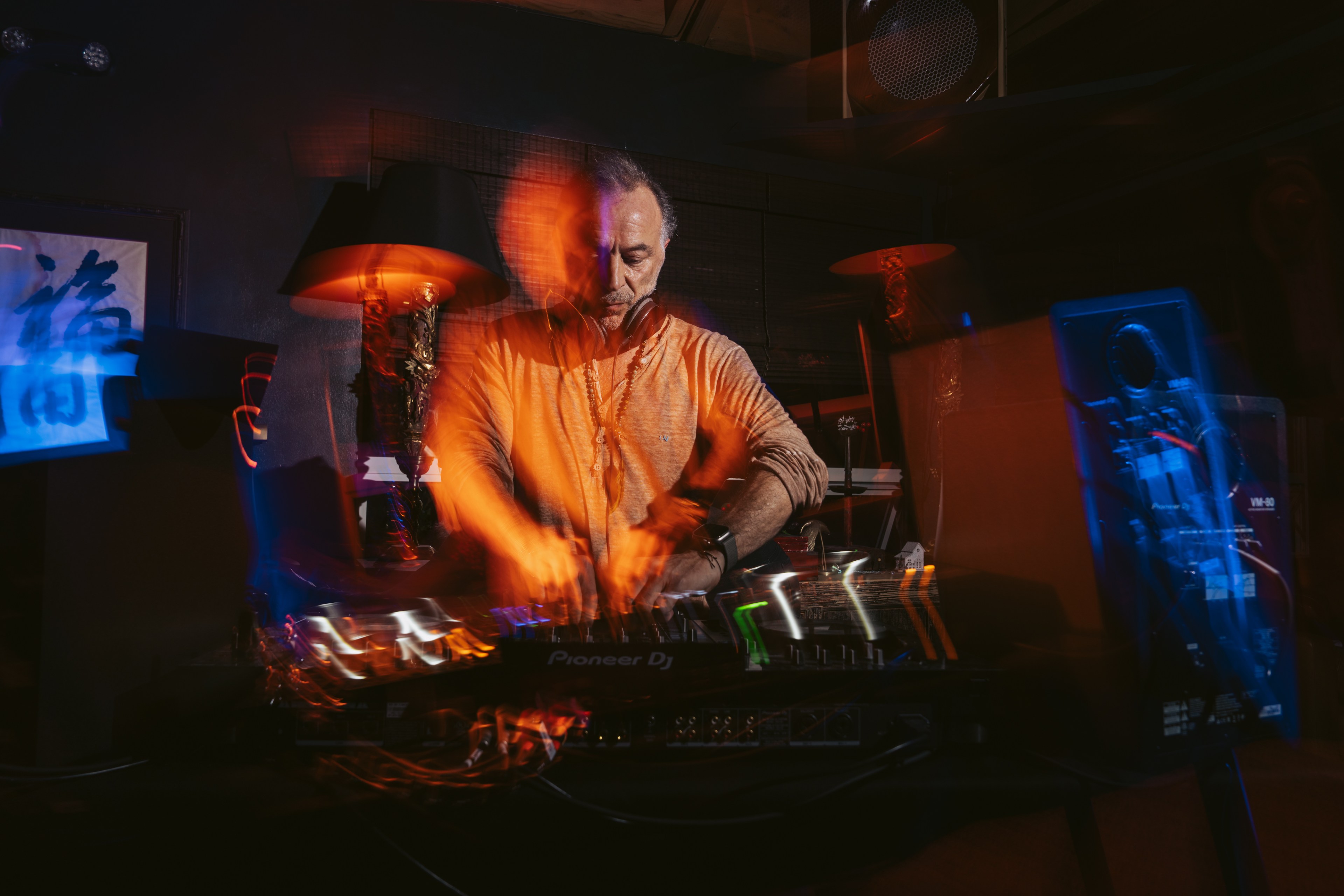 A DJ wearing headphones energetically works on a Pioneer DJ console in a dimly lit room, with motion blur and colorful lighting creating a dynamic atmosphere.