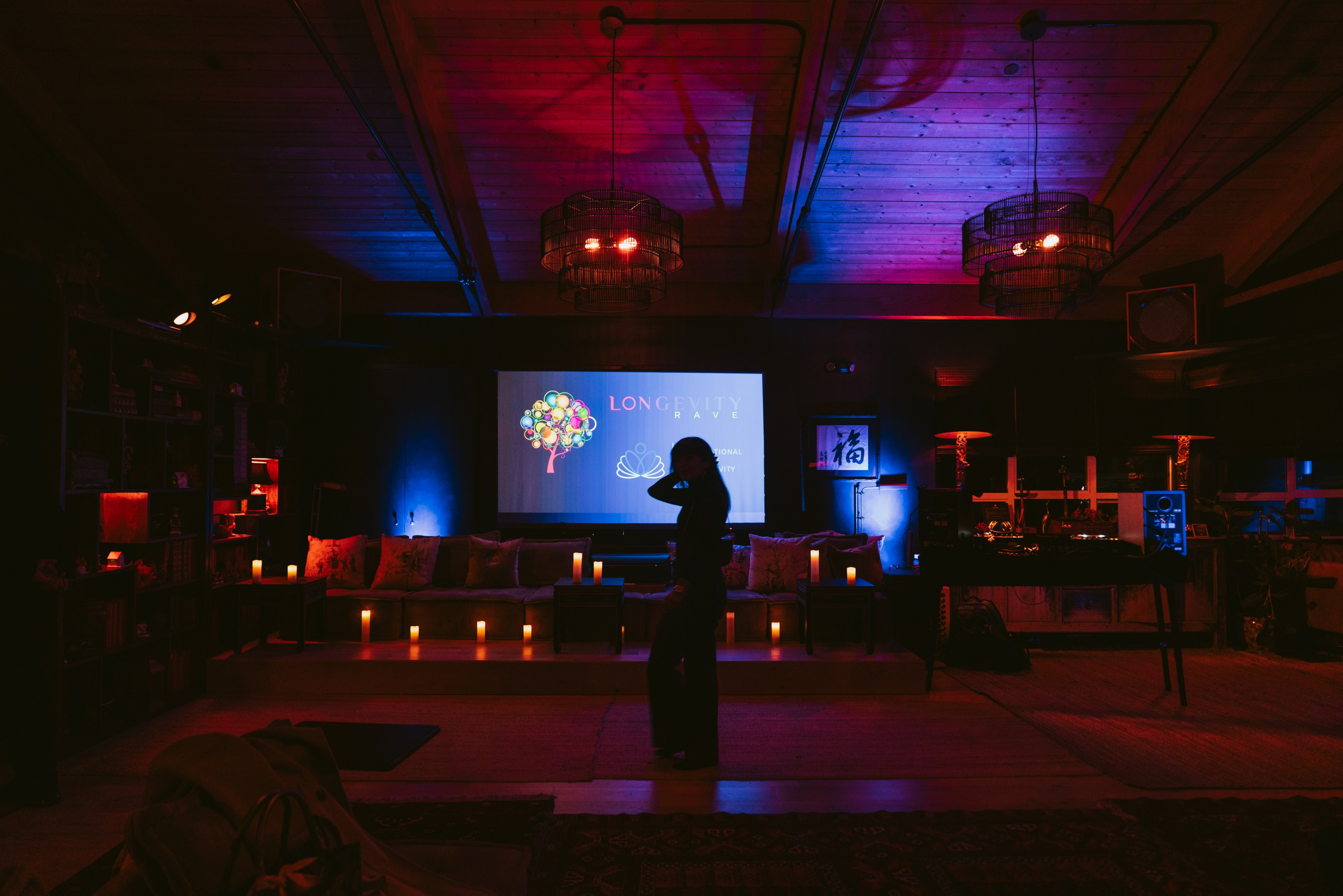 A dimly lit room has colorful lighting with a projector displaying &quot;LONGEVITY&quot; and an abstract tree. Candles line a cozy couch, and a person stands silhouetted.