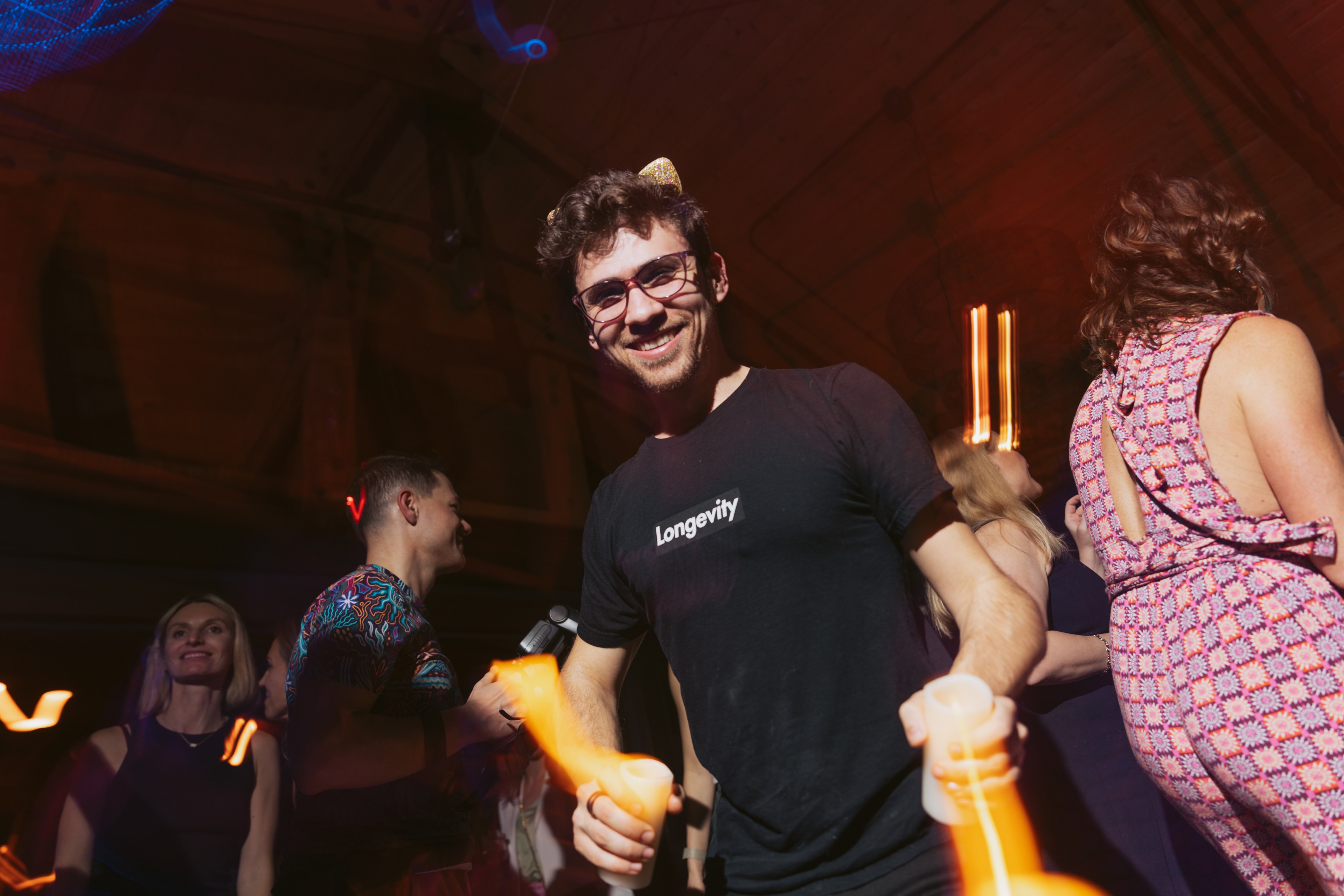 A smiling man in a &quot;Longevity&quot; T-shirt holds glowing cups at a party. Other people around him are dancing, with colorful lights enhancing the lively atmosphere.