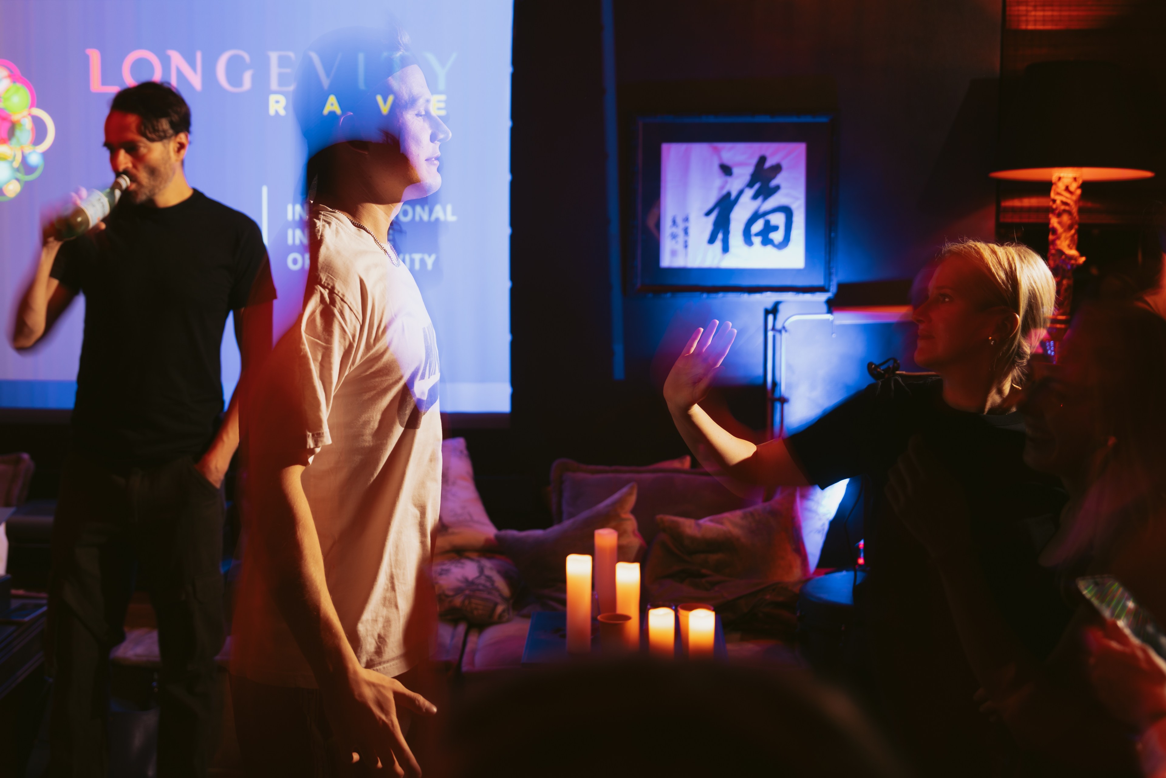 A dimly lit room with a projector displaying &quot;LONGEVITY RAVE&quot;. Three people are interacting, with candles on a table and a framed piece of art on the wall.