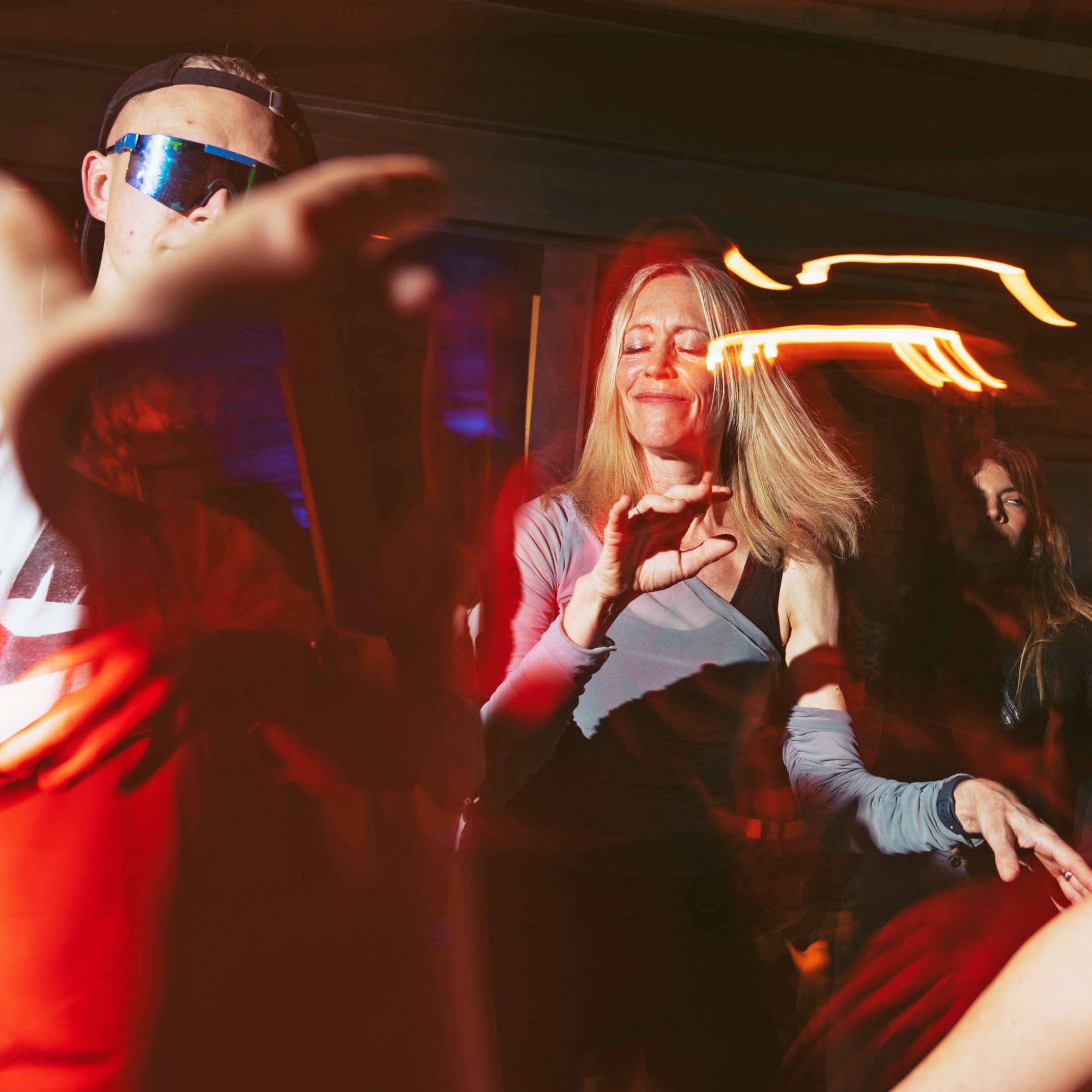 People are dancing energetically in a dimly lit space with colorful lights. A woman in the center smiles with her eyes closed, surrounded by blurred motion.