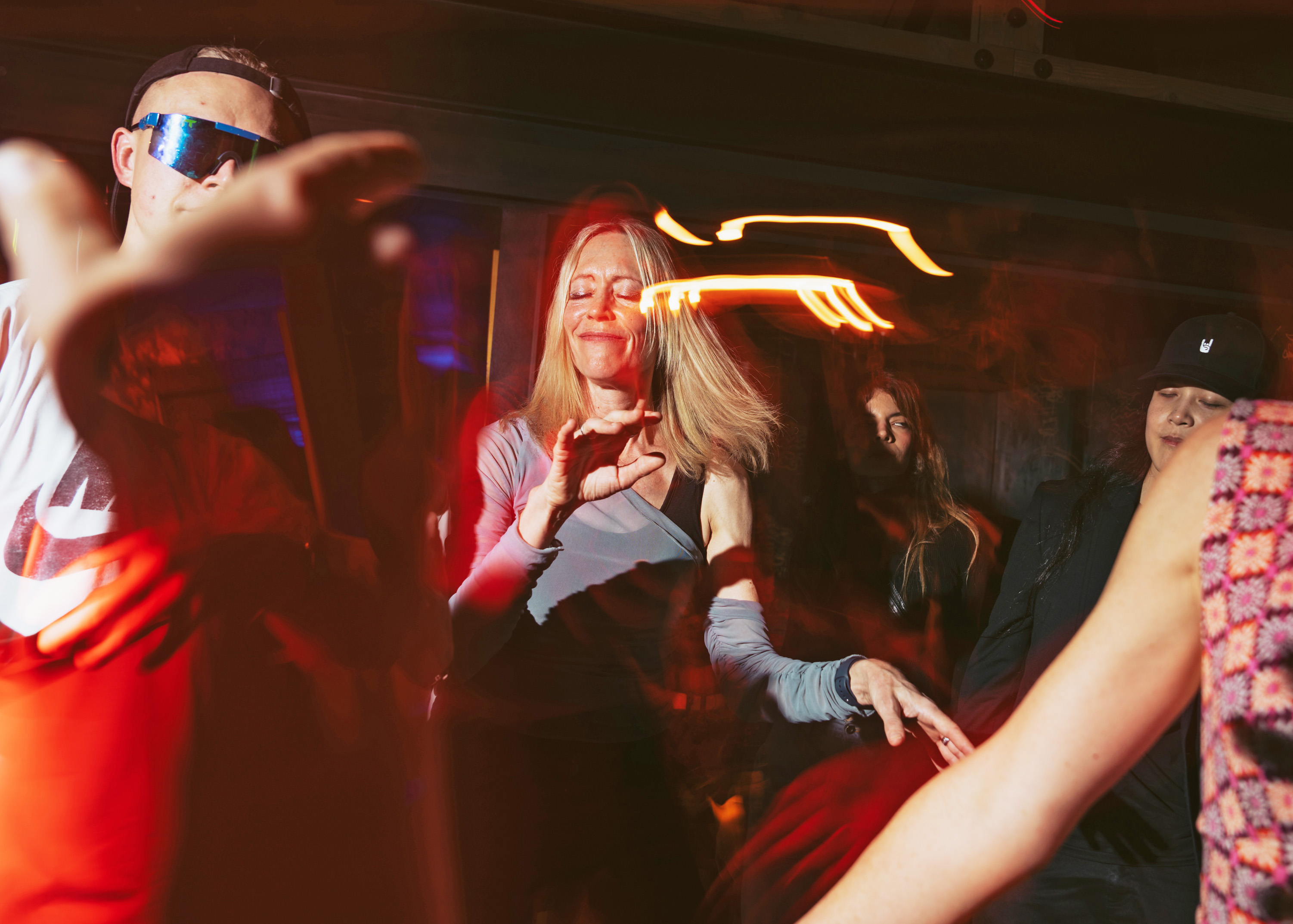 People are dancing energetically in a dimly lit space with colorful lights. A woman in the center smiles with her eyes closed, surrounded by blurred motion.