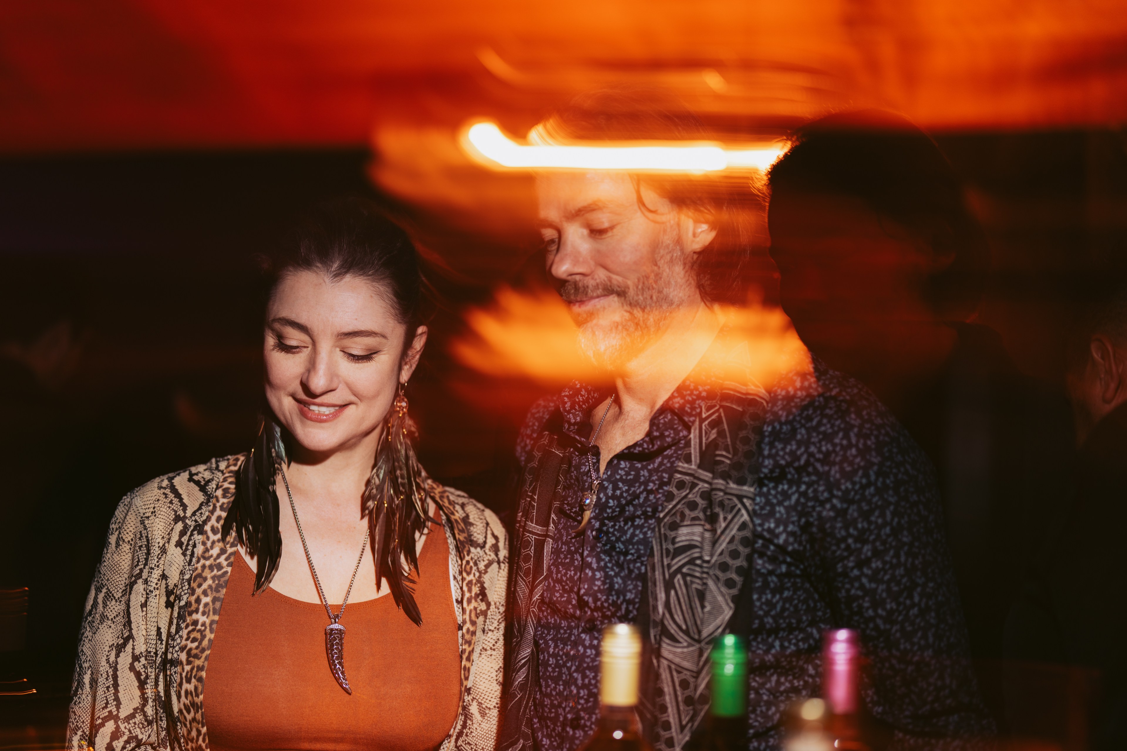 A woman and man stand together, softly lit in warm orange light. They appear relaxed, with blurred motion effects creating a dreamy atmosphere.