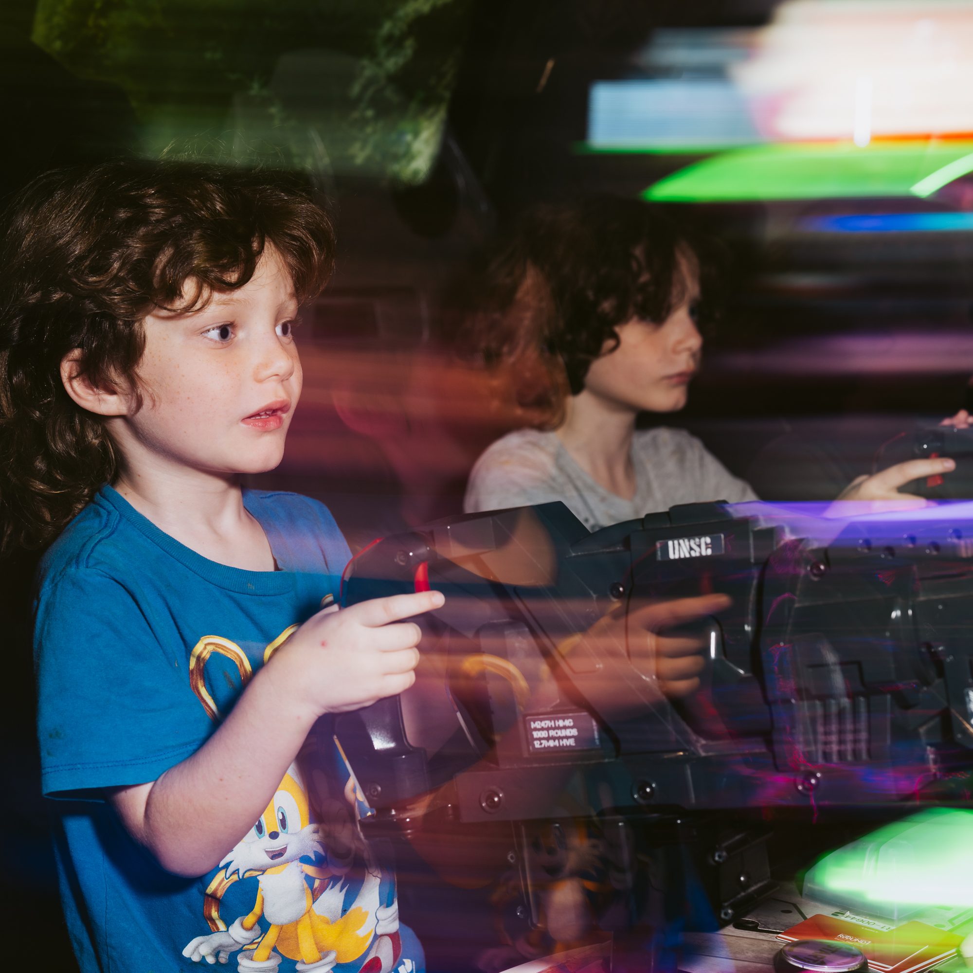 Two children are playing an arcade shooting game. One child in a blue Sonic t-shirt is in focus, holding a gun controller, with colorful lights around them.