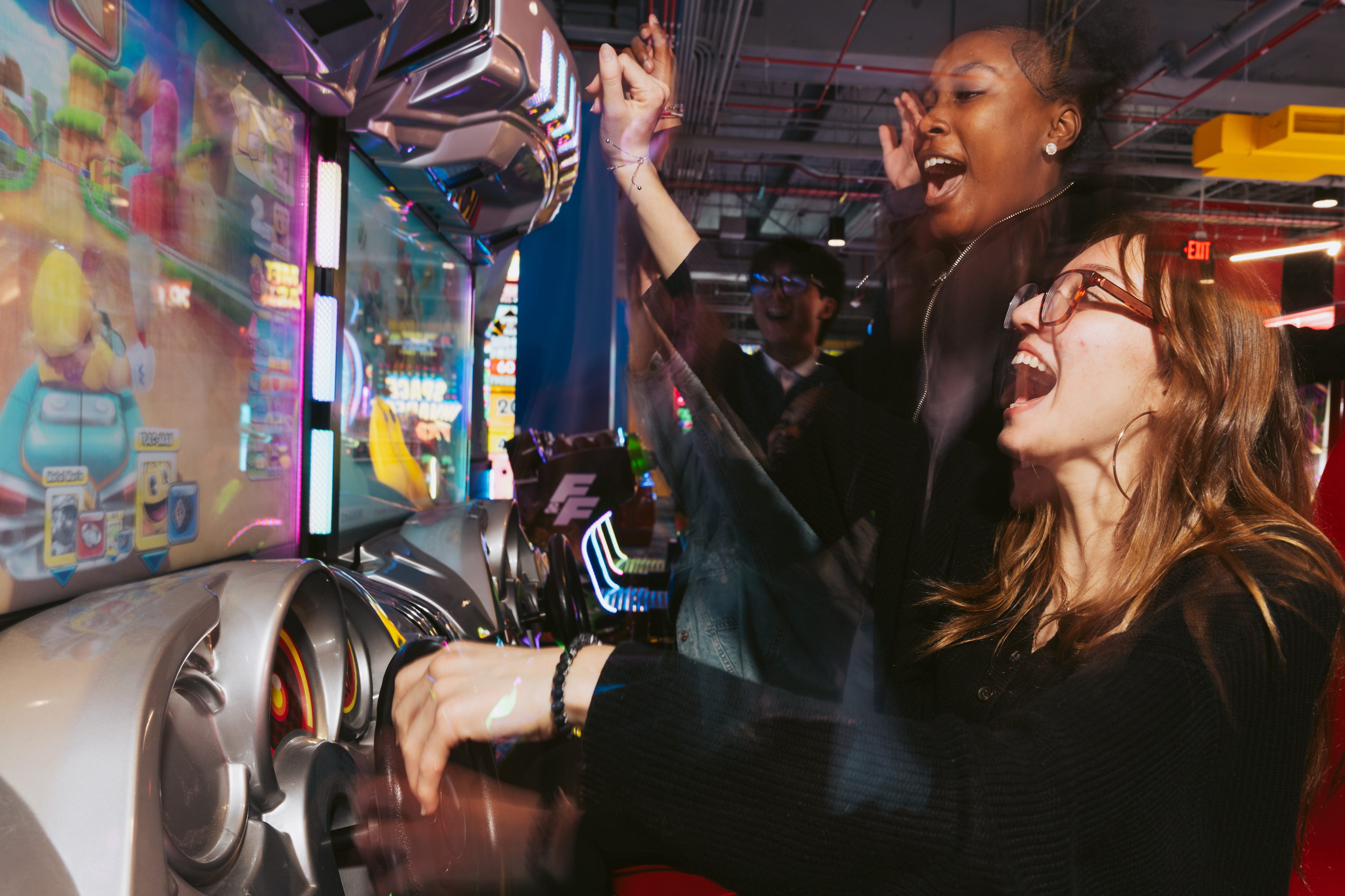 Two excited people play an arcade racing game, cheering with raised arms. Bright screens and vivid colors surround them, capturing a lively atmosphere.