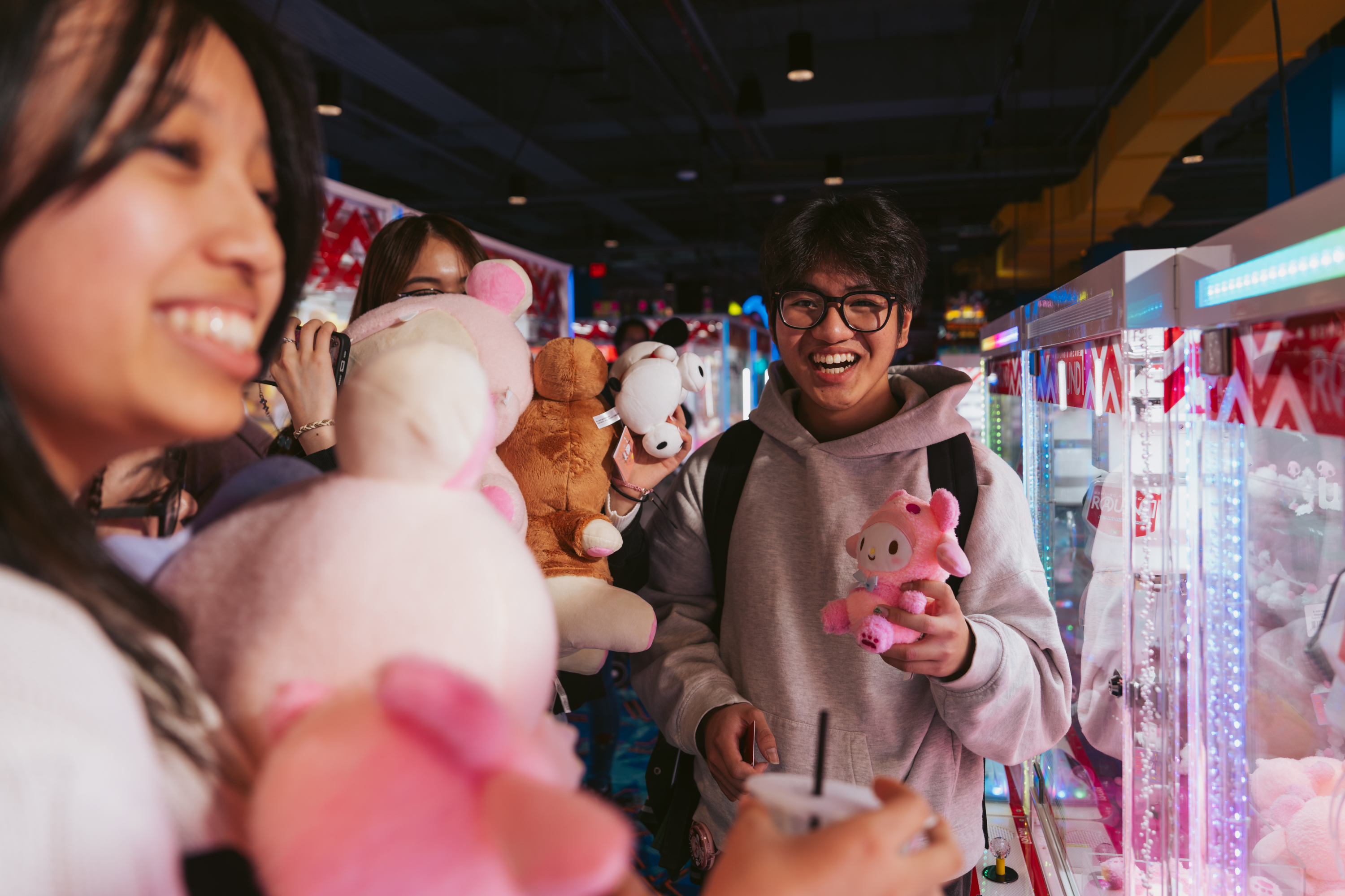 A group of smiling people in an arcade holds plush toys near a claw machine; one person in a hoodie holds a drink and a small pink plush.