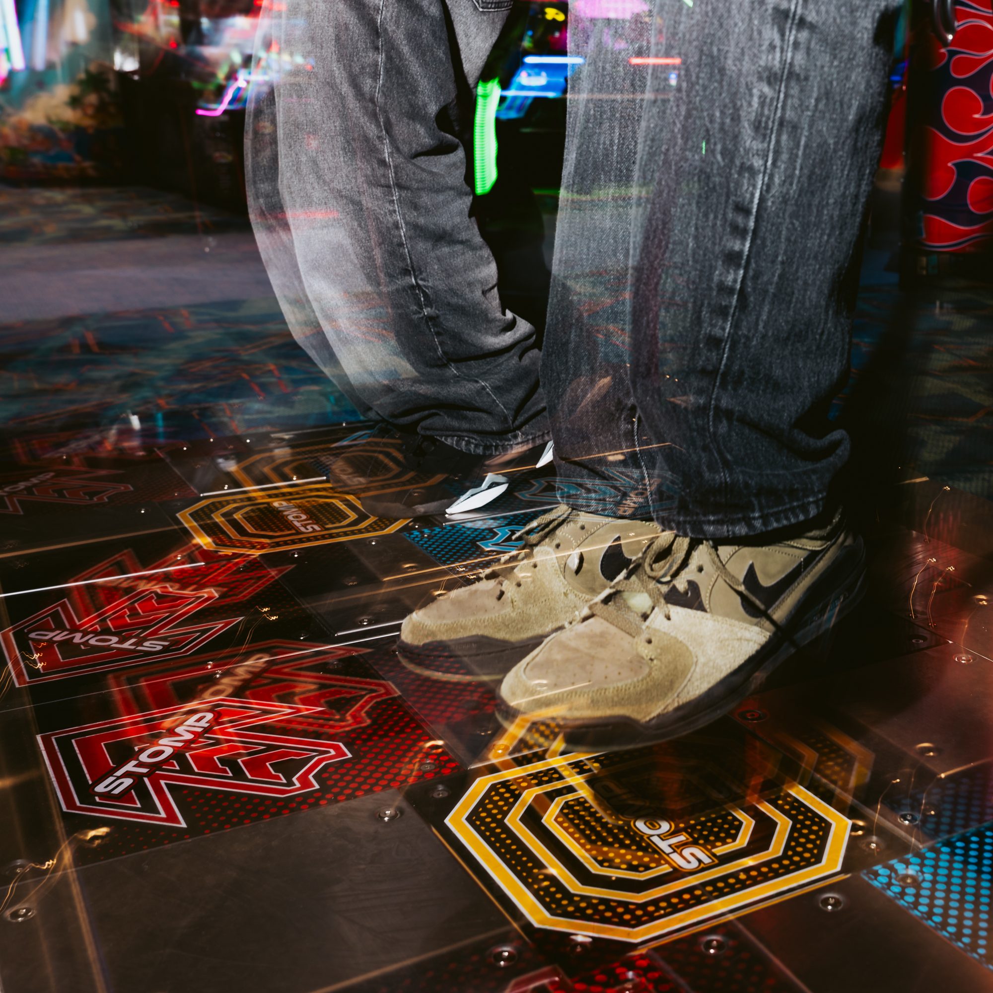 A person wearing green Nike sneakers and dark jeans stands on a colorful arcade dance machine platform, with red railings and bright patterns visible.