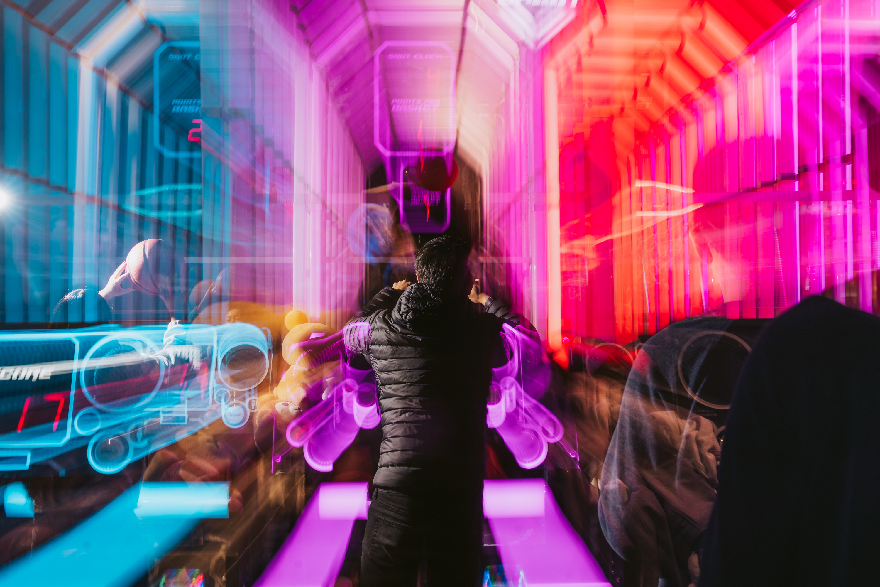 The image shows a person in a dark jacket standing in a vibrant, futuristic arcade with bright pink and blue neon lights, creating a dynamic and colorful blur effect.