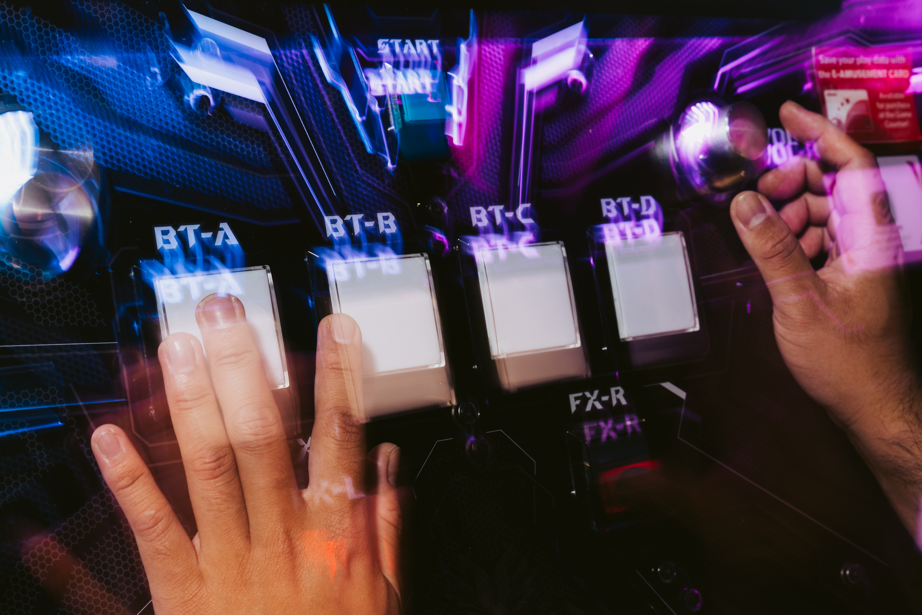 Two hands are interacting with a rhythm game machine, pressing large white buttons labeled BT-A to BT-D and FX-R, under vibrant blue and pink lighting.