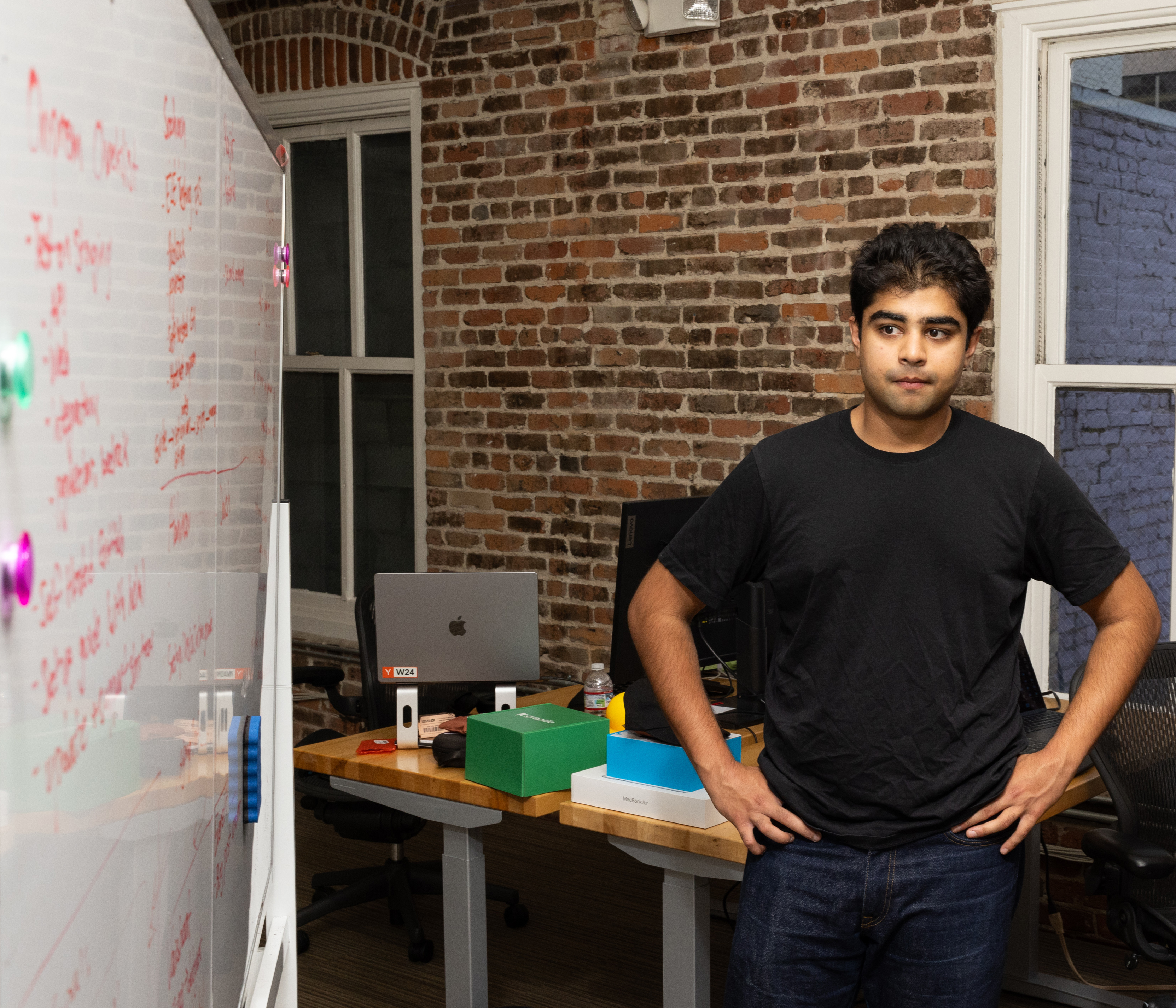 A person in a black shirt stands with hands on hips in an office with a brick wall, whiteboard, and desk holding laptops and colorful boxes.