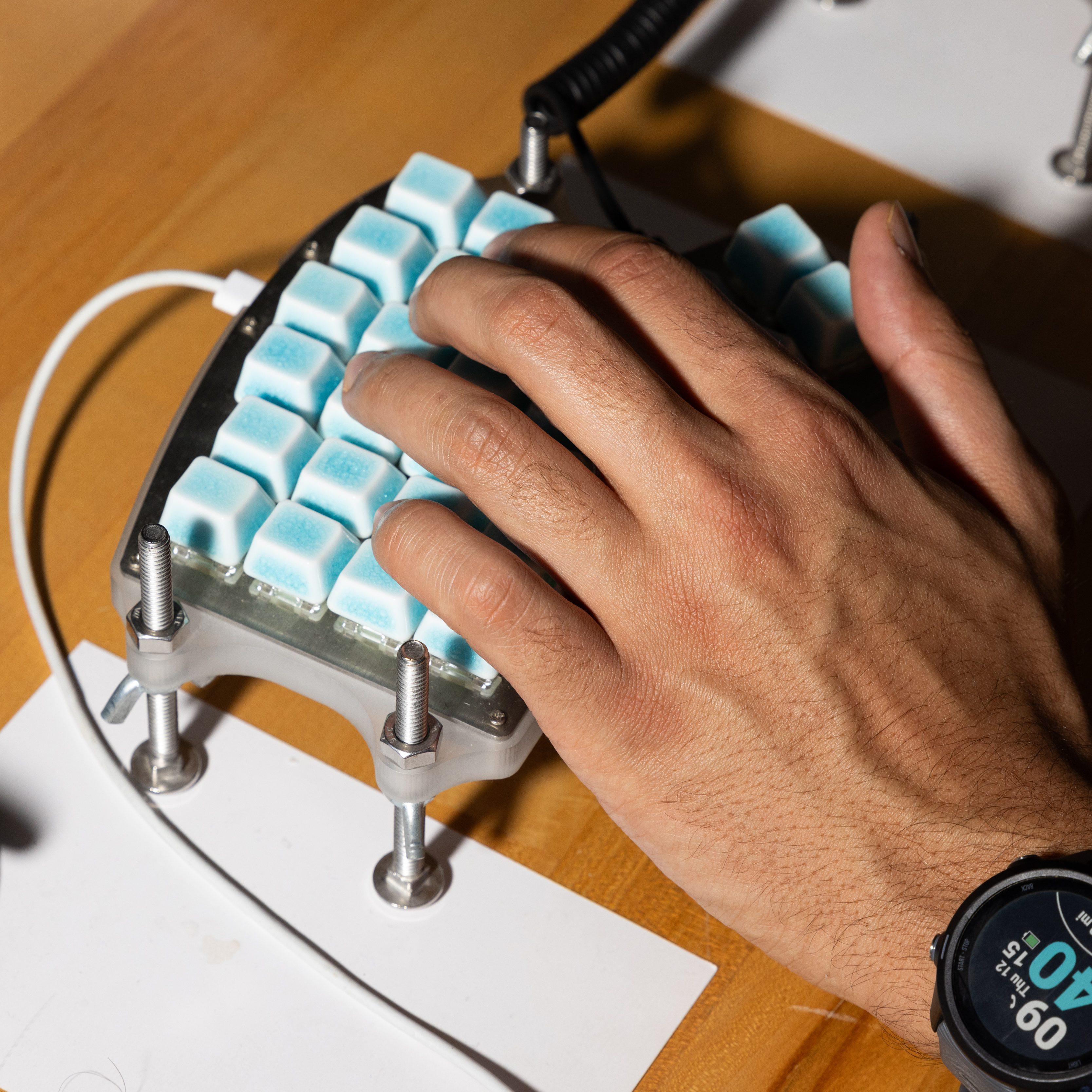 A hand is typing on a mechanical keyboard with light blue keycaps. The keyboard has a unique, raised design secured with metal bolts. A smartwatch is visible.