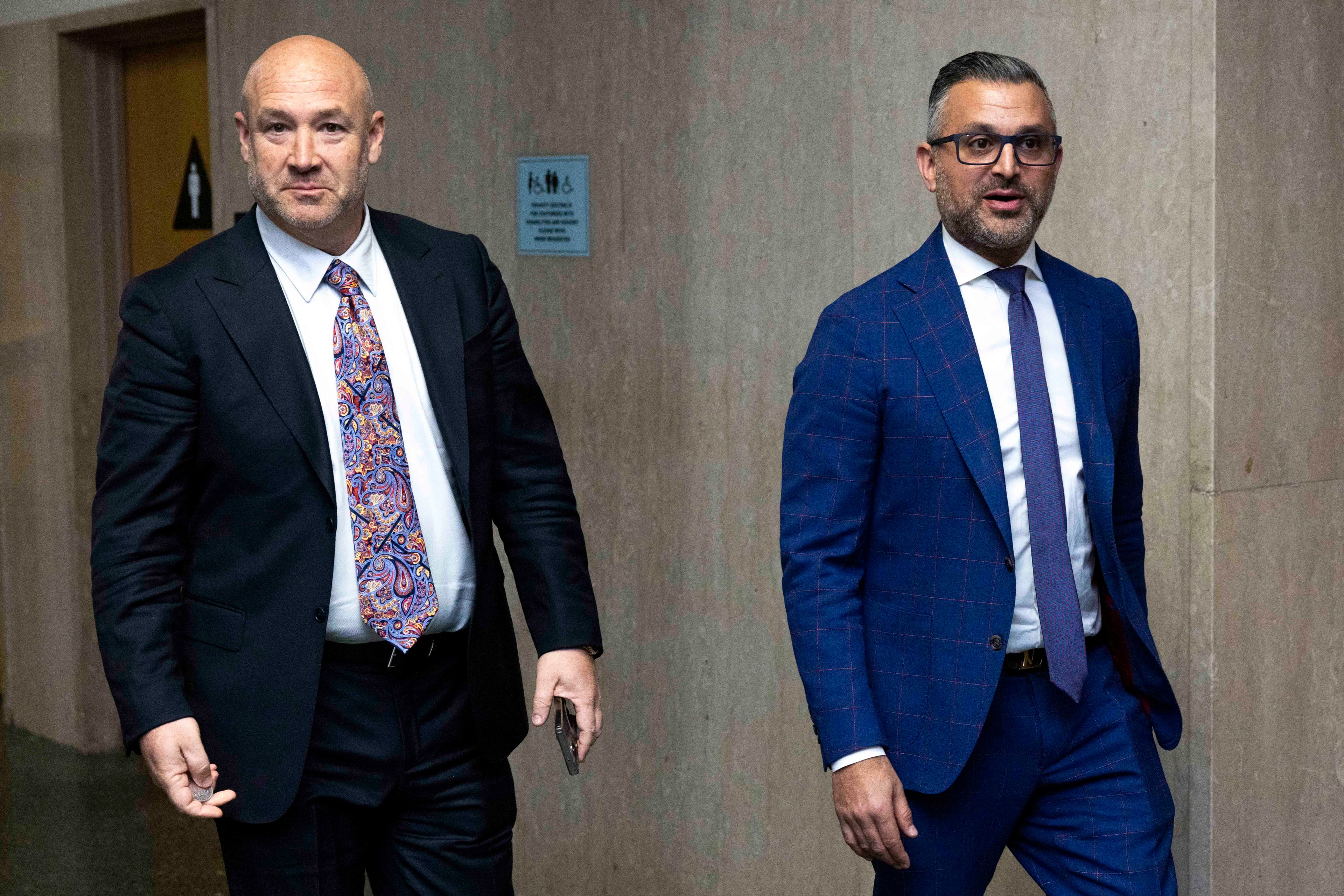 Two men in suits walk down a hallway. The man on the left wears a black suit with a colorful tie, while the one on the right wears a blue checkered suit.