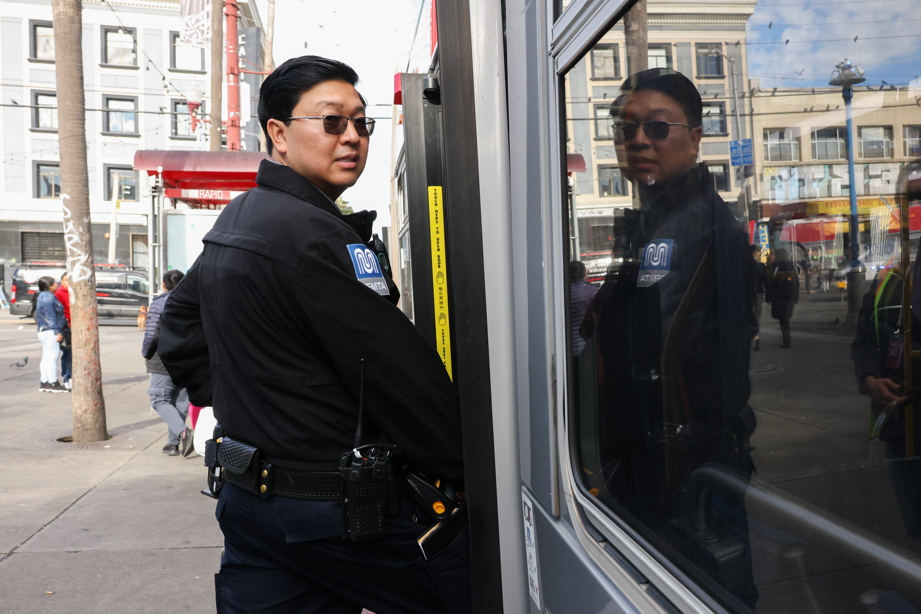 A uniformed security officer stands near a bus, with their reflection visible in its window, on a busy city street with pedestrians and buildings in the background.