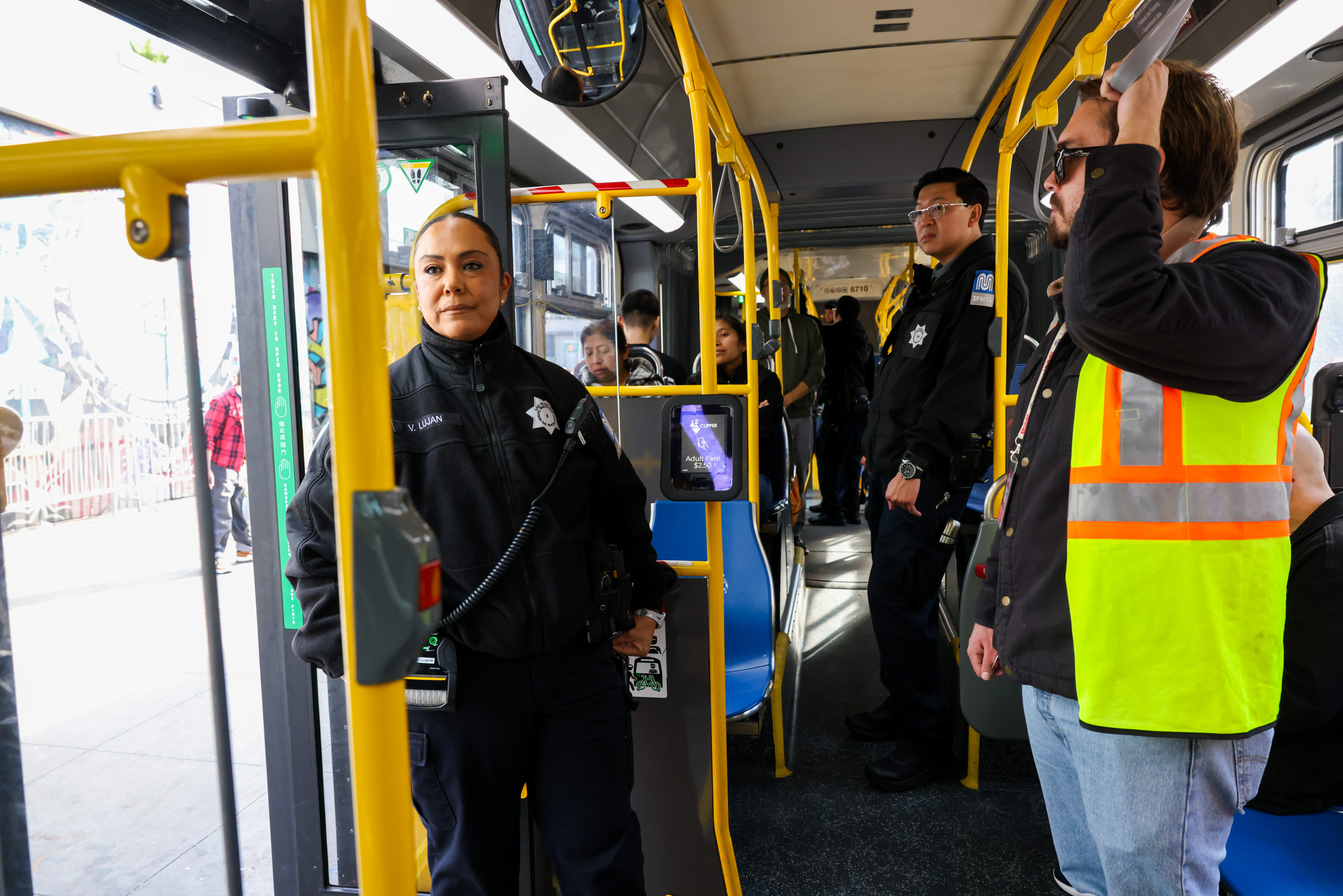 A female officer in uniform stands near a bus door, with another officer and a man in a reflective vest inside. Passengers sit and stand throughout the bus.