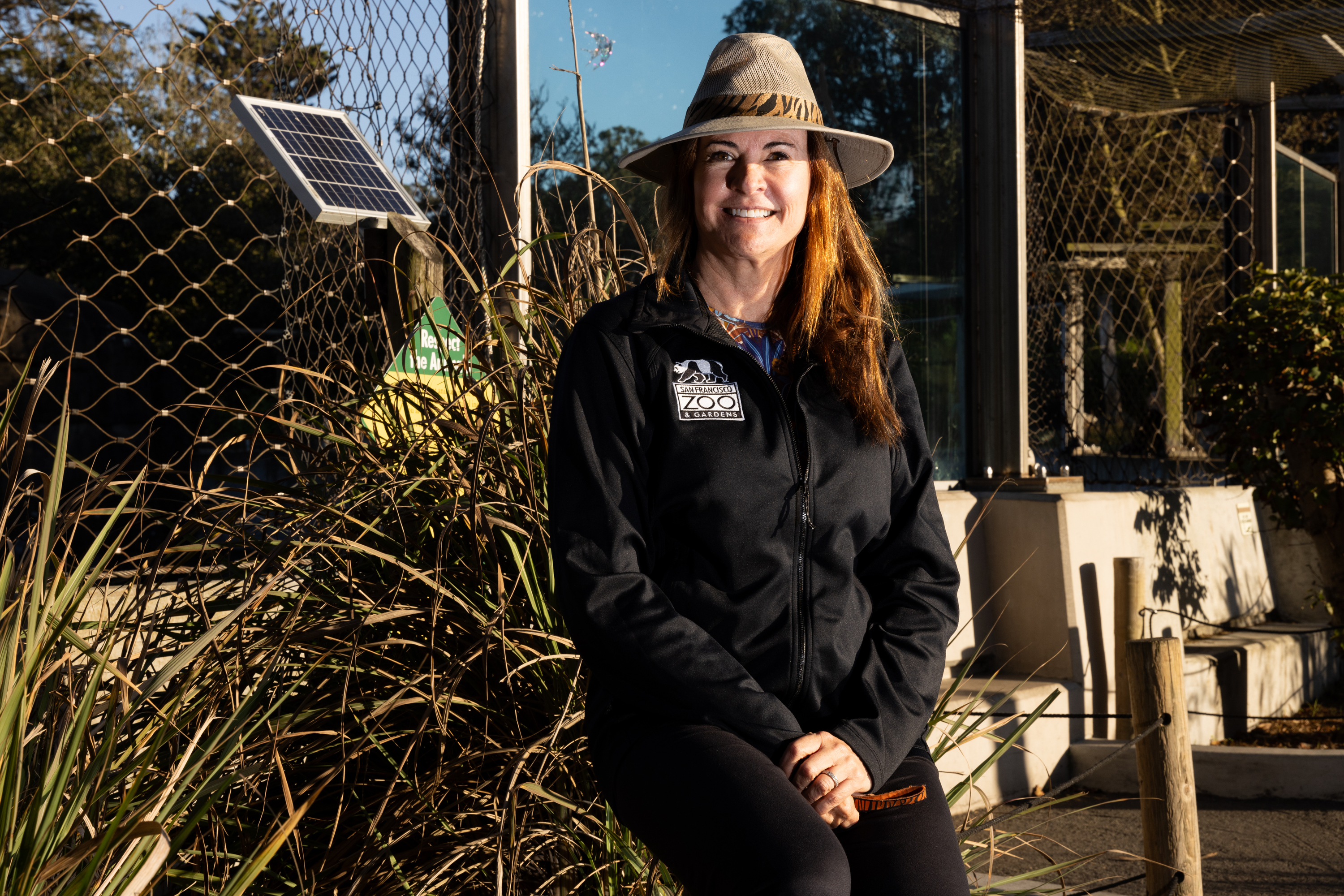 A woman in a safari hat and black zoo jacket smiles while sitting outdoors near a wire fence, with plants and a small solar panel in the background.