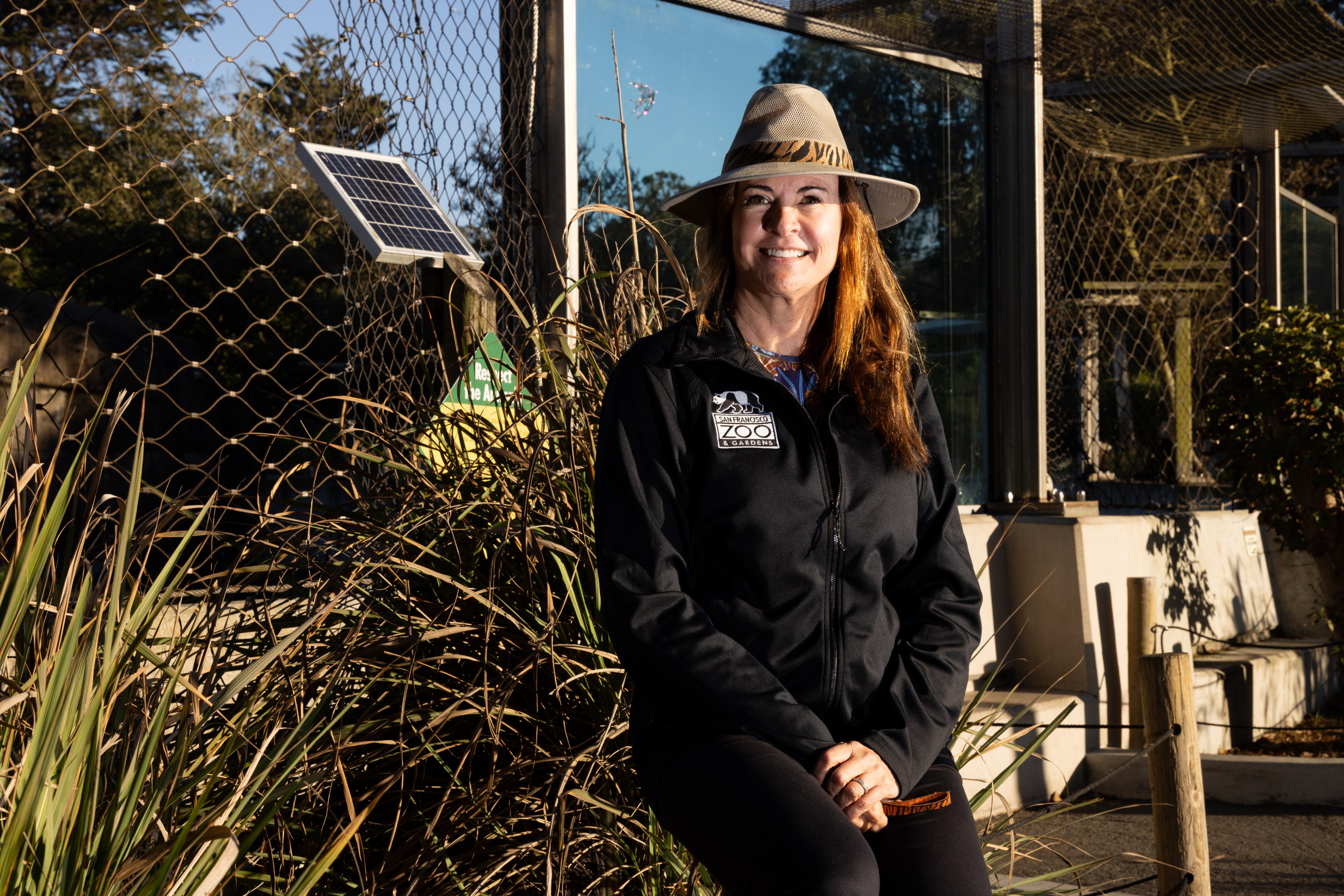 A person wearing a wide-brimmed hat and a black jacket with a zoo logo stands smiling beside a mesh enclosure and solar panel with foliage around.