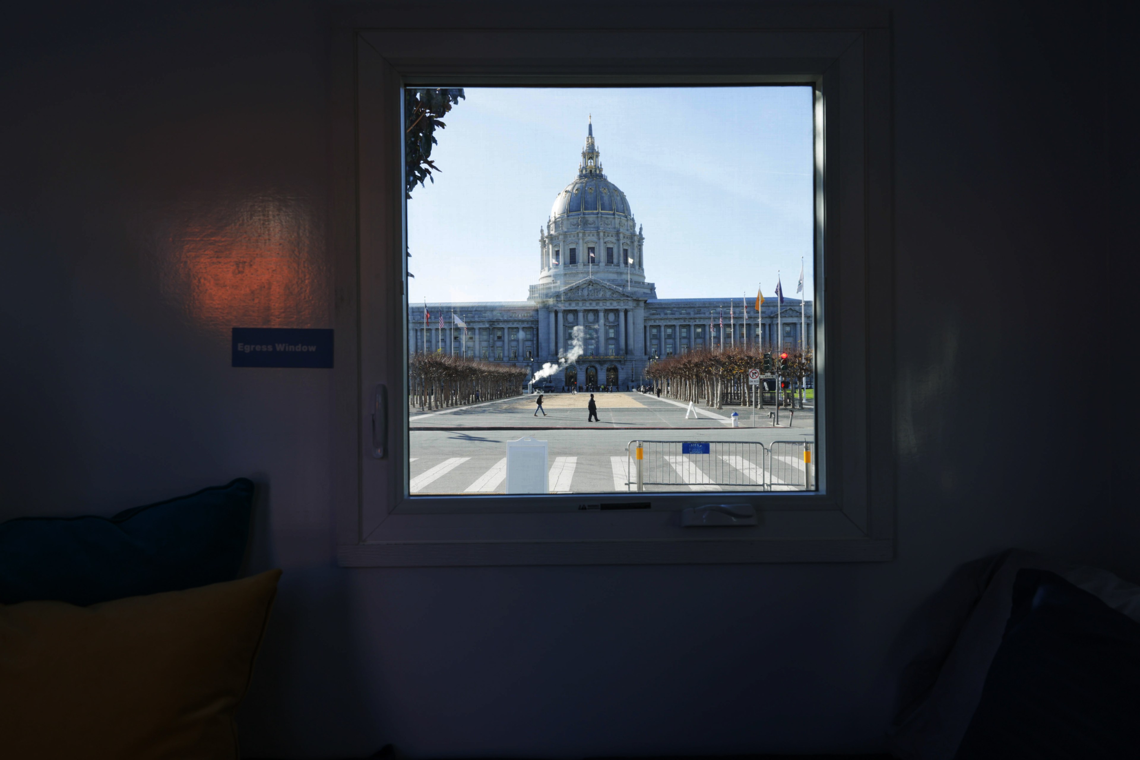 A grand building with a domed roof is framed through a window. Flags line the path leading to it, and a couple of people walk across the plaza.